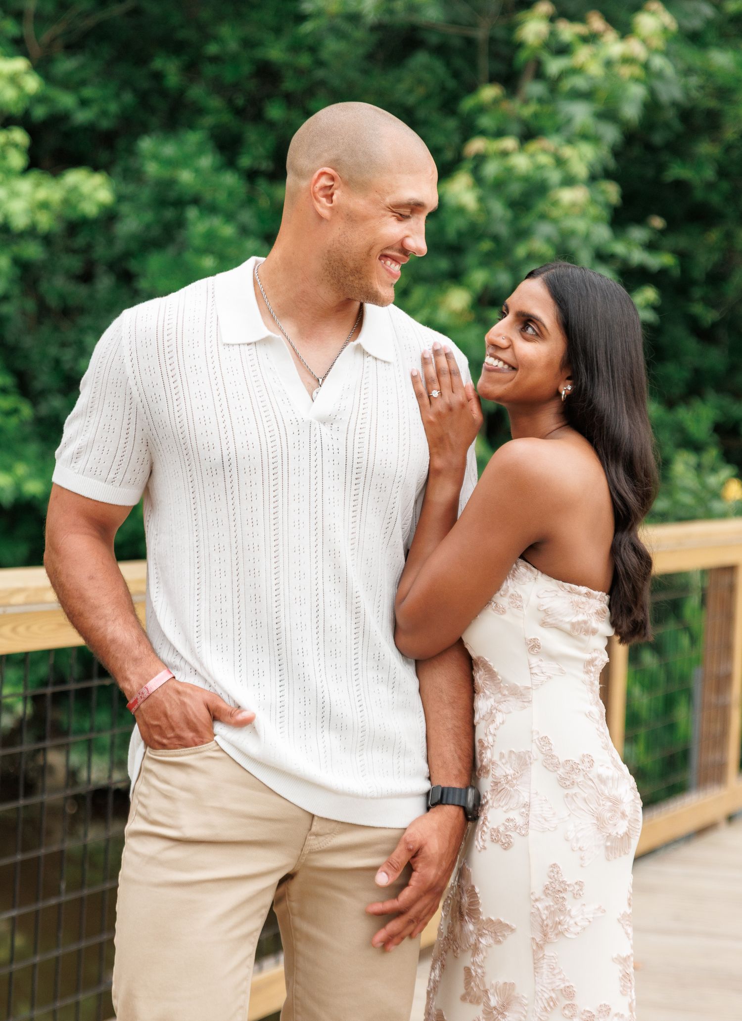 A smiling couple poses on a wooden bridge surrounded by greenery, woman in floral dress touching man's white polo shirt.
