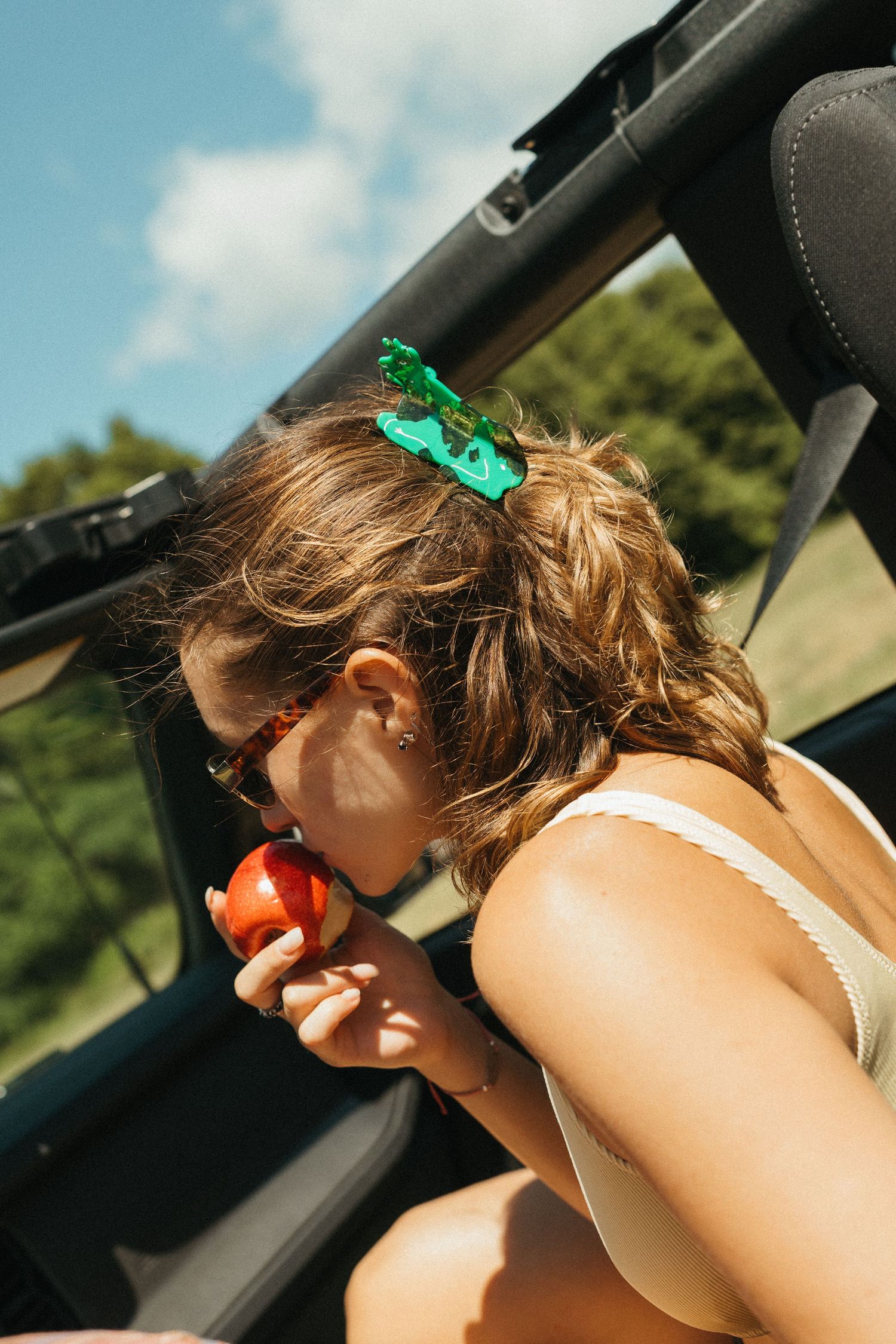 Woman in white top eating a red apple in a convertible car on a sunny day.
