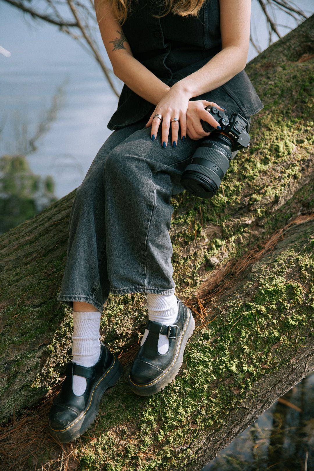 Woman in gray jeans and black shoes sitting on mossy log holding a camera.