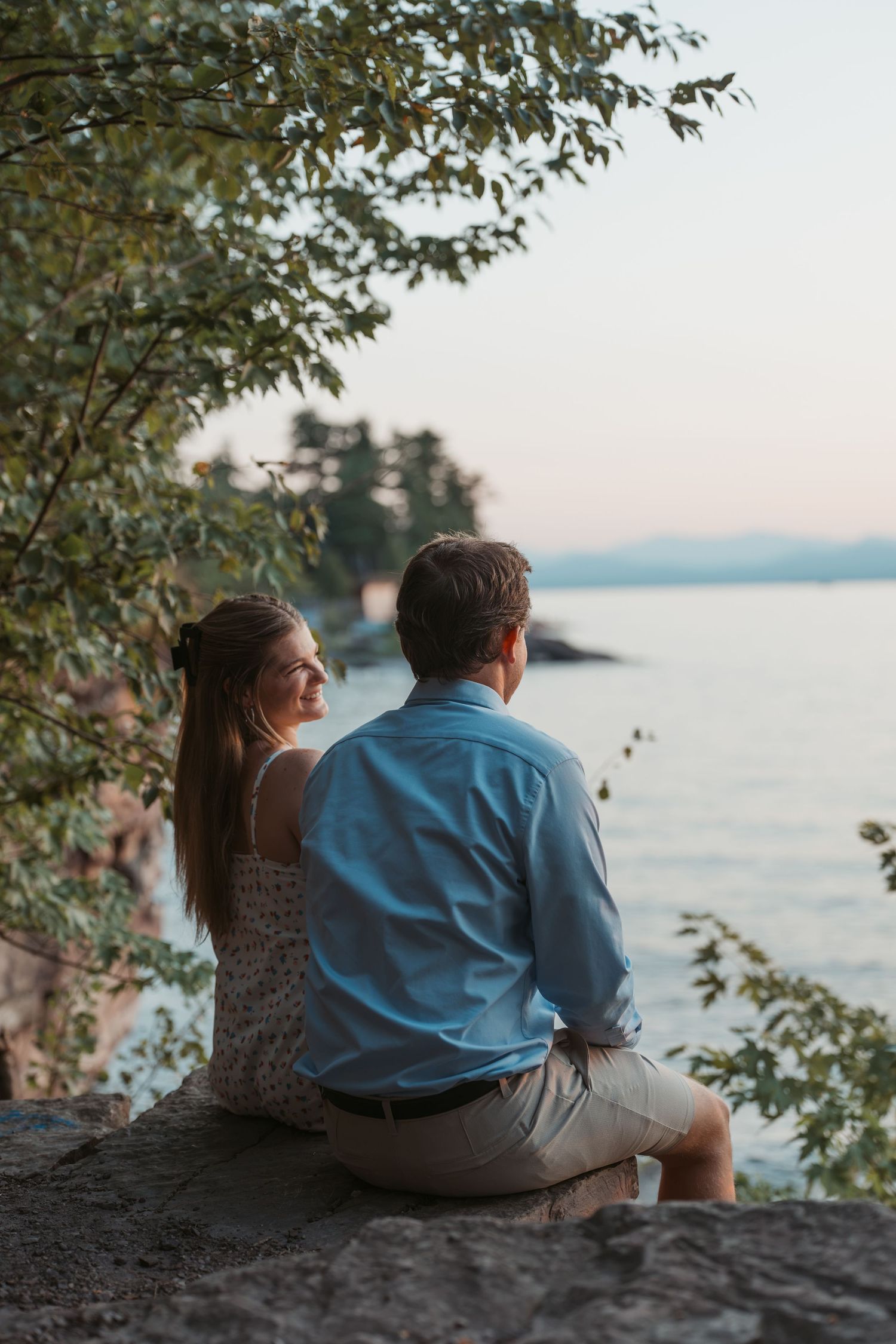 Couple sitting on rocks overlooking a serene lake at dusk under tree branches.