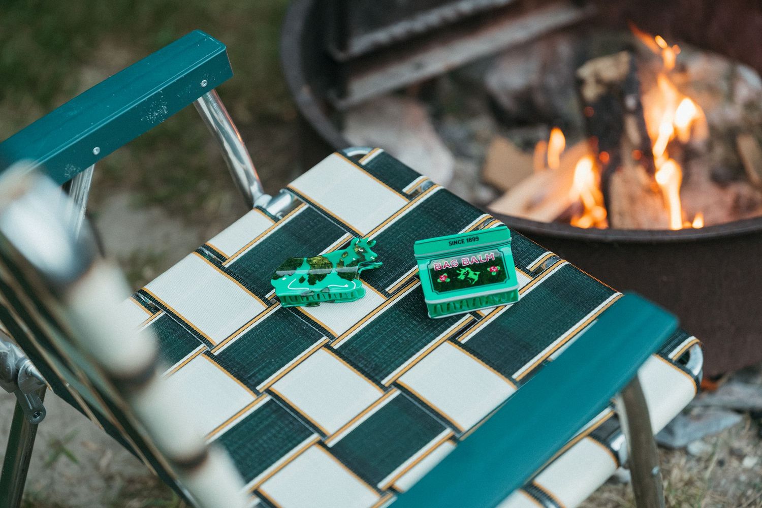 Two green Bag Balm hair clips on a woven camp chair near a fire pit.