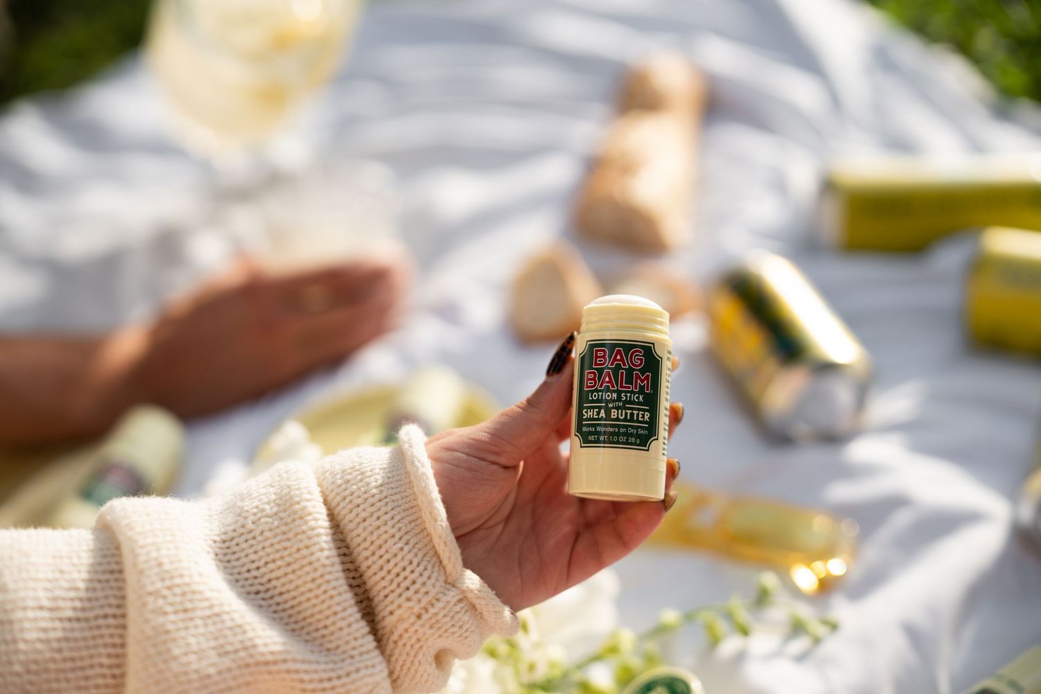 Hand holding Bag Balm stick over white picnic blanket with LaCroix cans.
