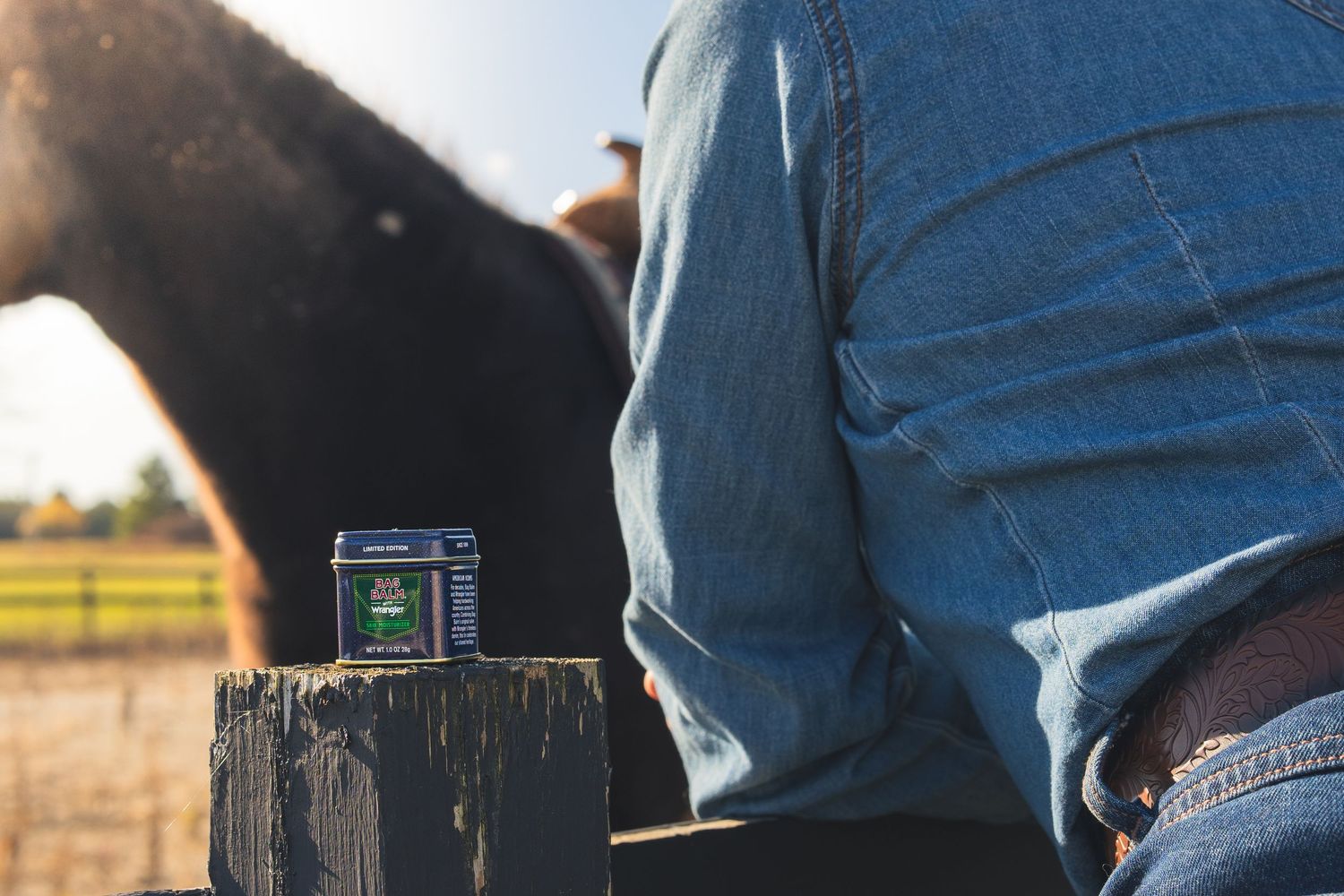 Bag Balm x Wrangler limited edition tin on fence post beside person in jeans near horse.