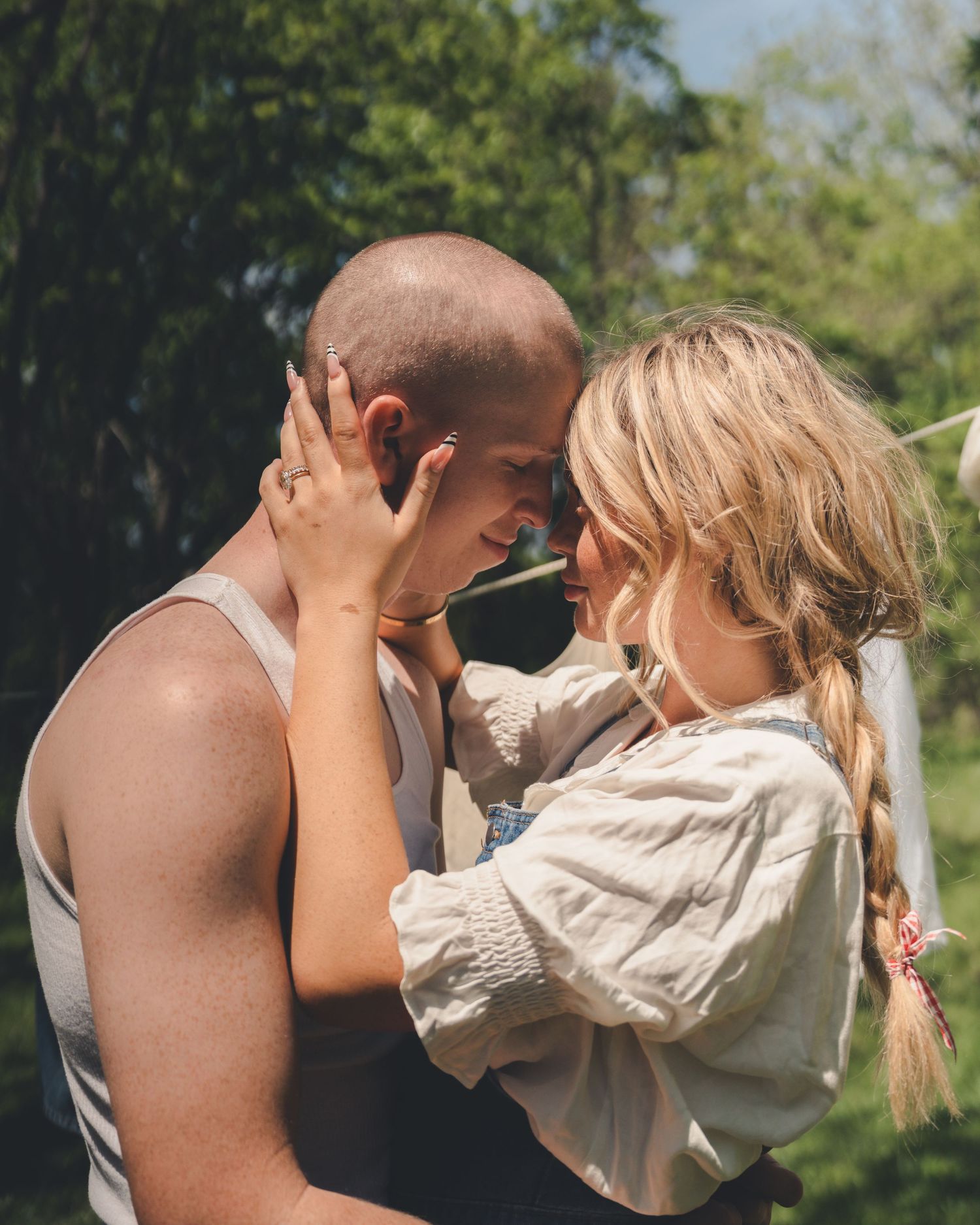 Romantic couple leaning foreheads together outdoors surrounded by green trees on a sunny day.