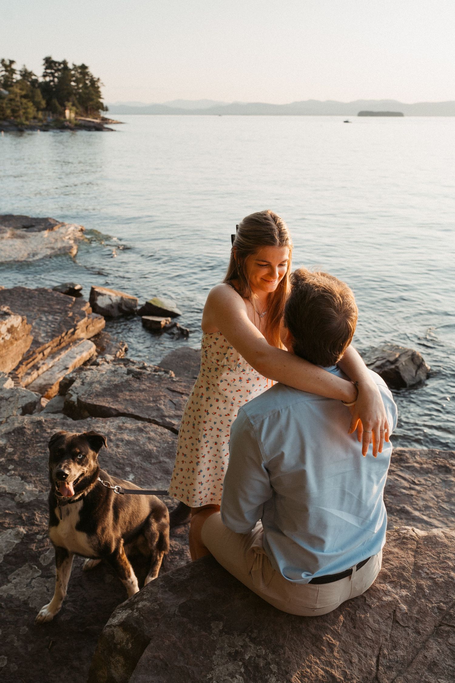 A couple embraces on rocky shoreline at sunset with their dog beside them and calm lake views.