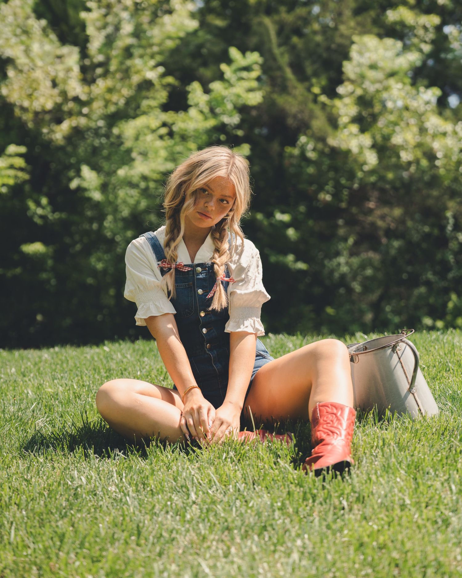 Young woman in denim overalls and red cowboy boots sitting on grass in sunlight.