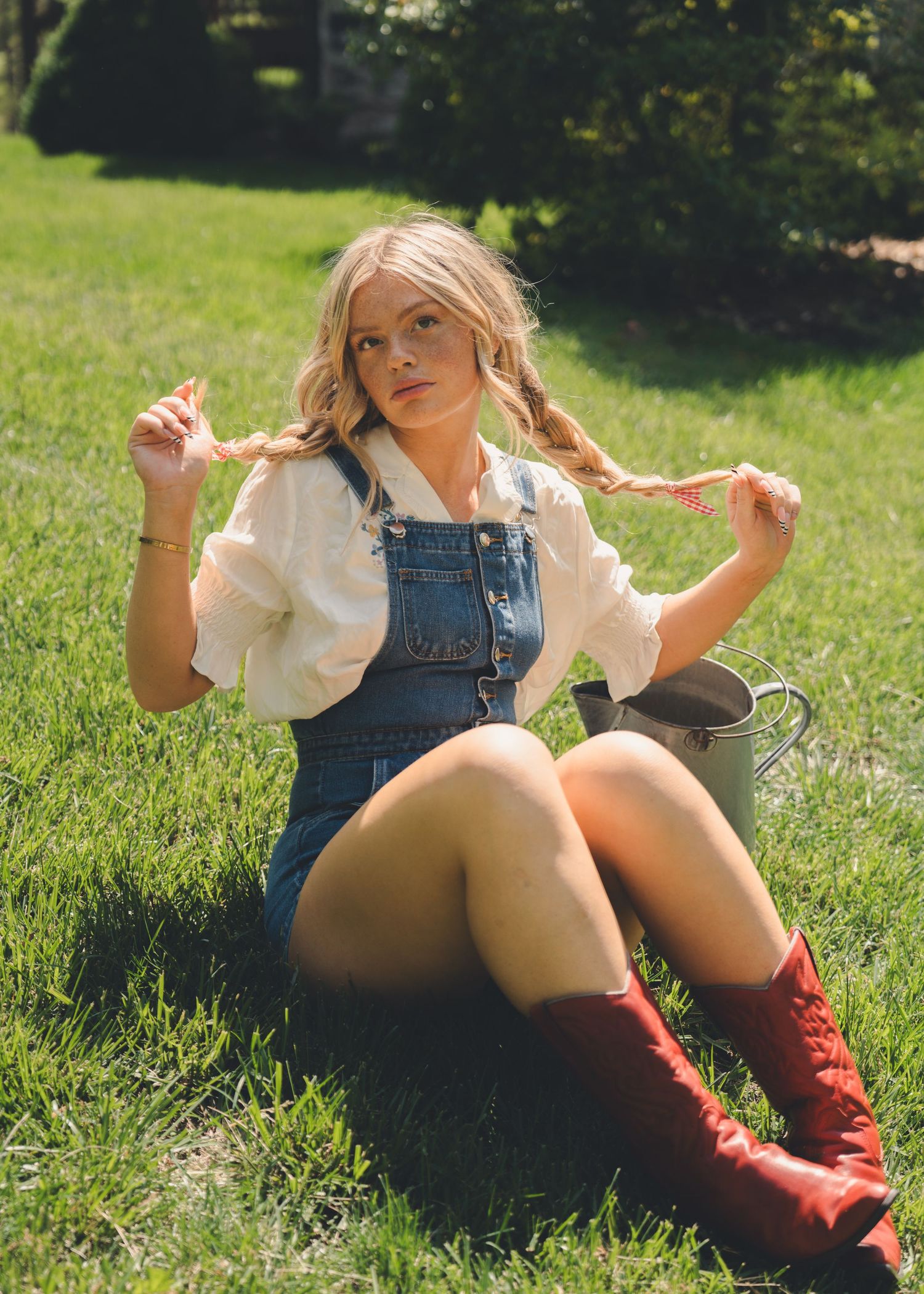 Young woman in denim overalls pulling her braids while sitting on green grass.