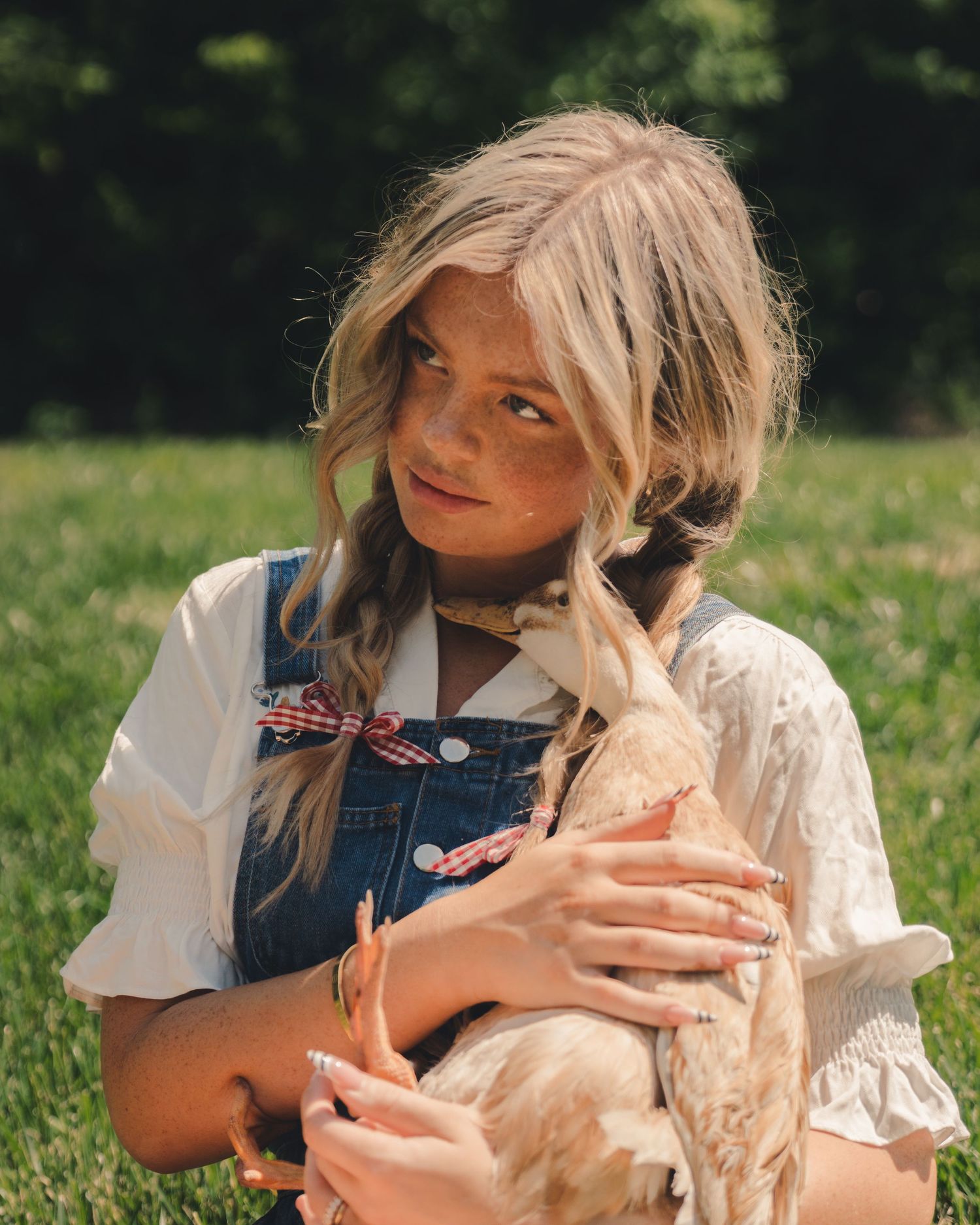 Young blonde girl holding a duck while sitting on green grass in overalls.