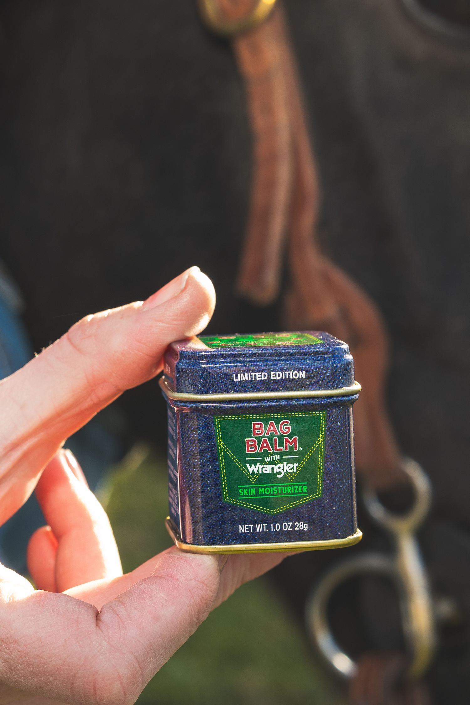 Hand holding limited edition Bag Balm x Wrangler skin moisturizer tin near a horse with bridle.