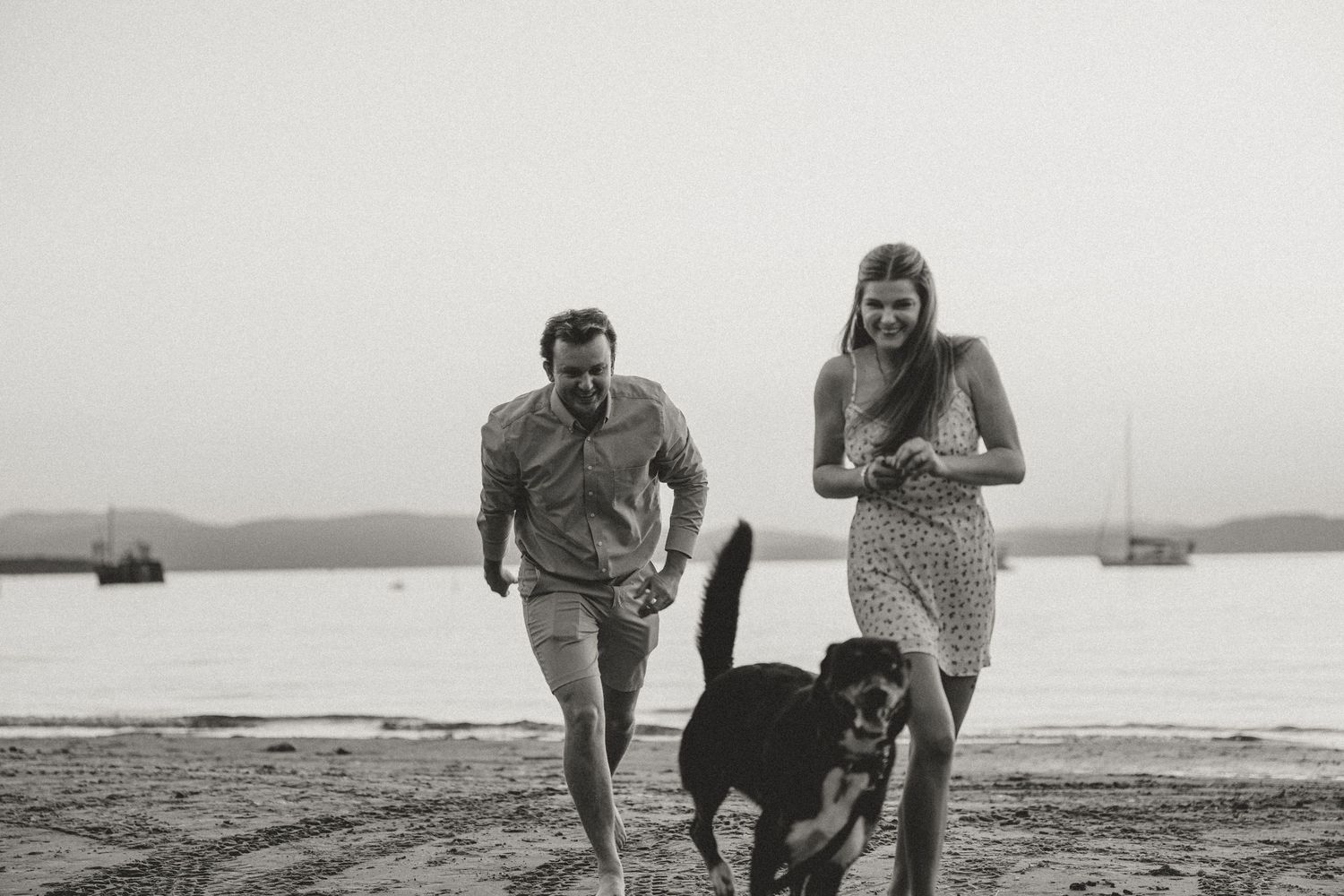 Couple running joyfully with their dog on a beach in black and white photo with sailboats in background.