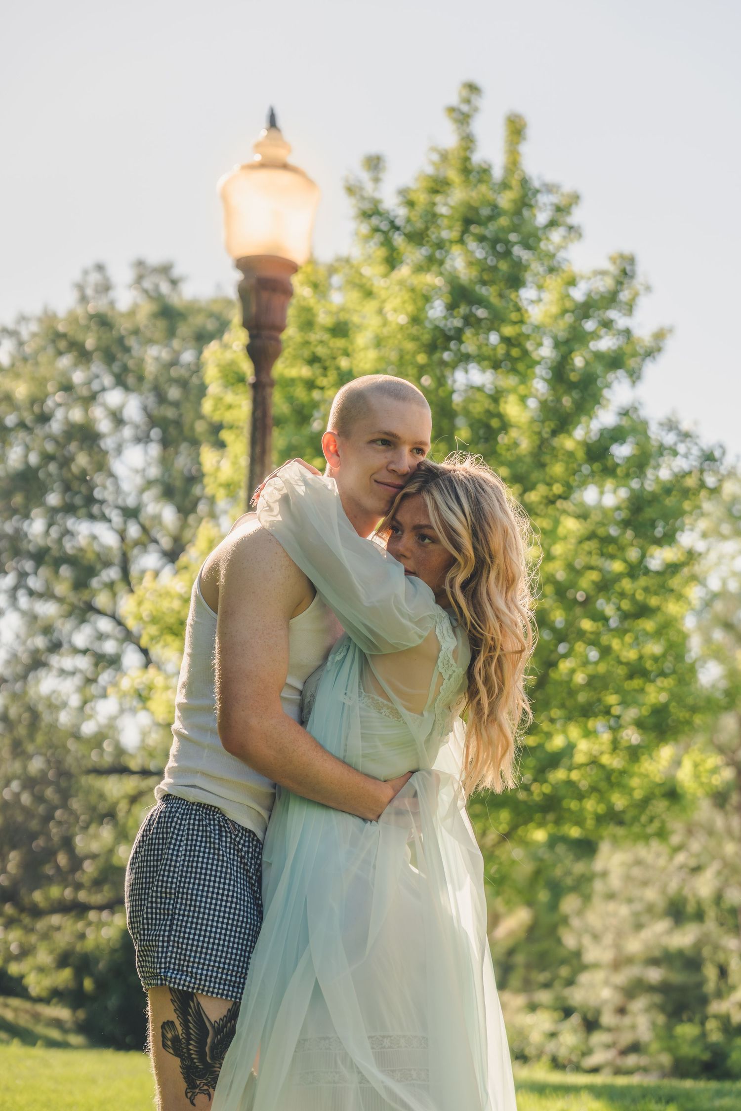 Couple embracing outdoors near a glowing street lamp surrounded by green trees in warm sunlight.