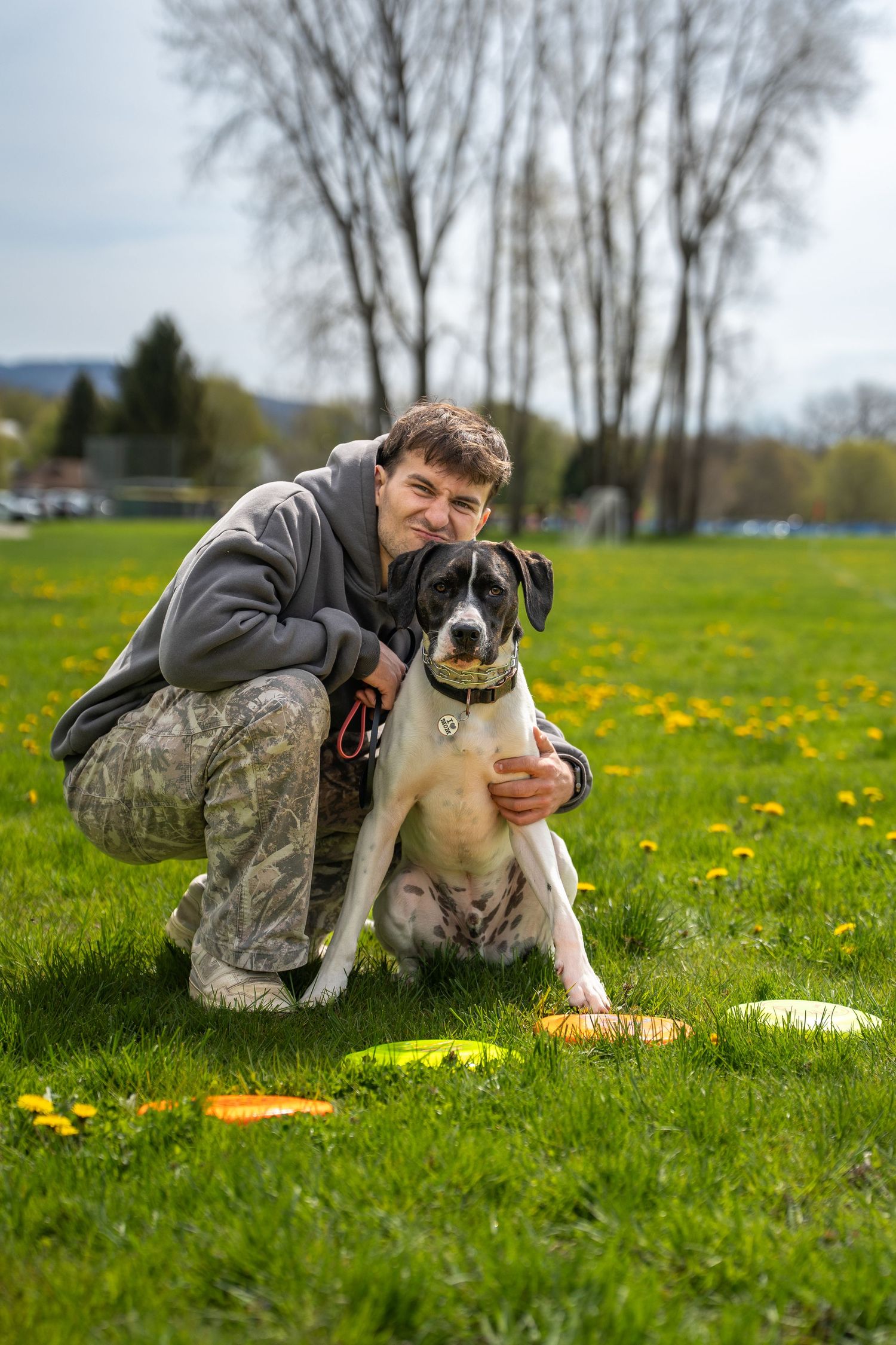 Man crouching with black and white dog on green grass field with yellow dandelions and frisbees nearby.