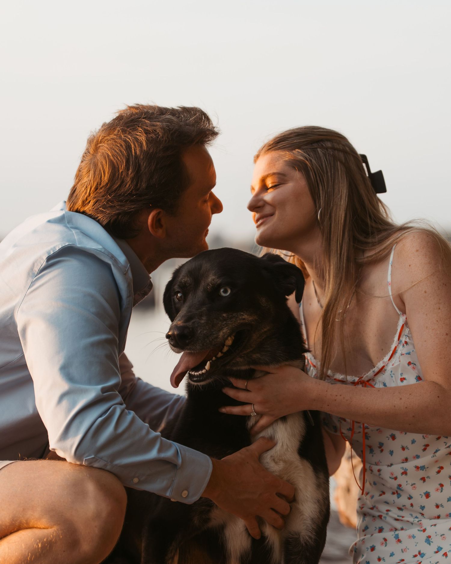 Couple smiling and cuddling with their happy black dog outdoors at sunset.