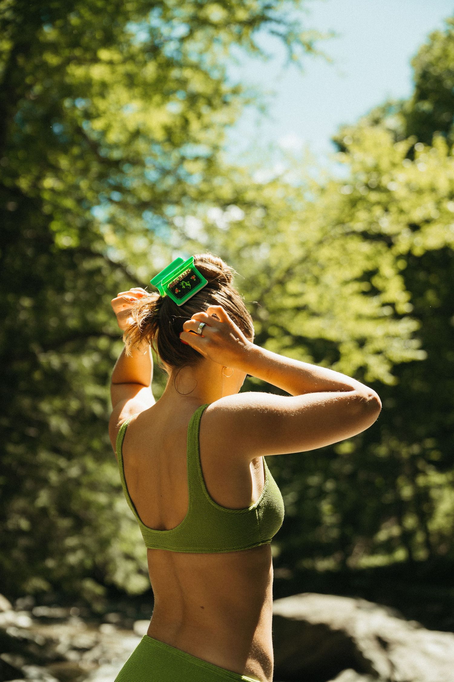 Woman in green sports bra placing a Bag Balm hair clip in her hair outdoors.