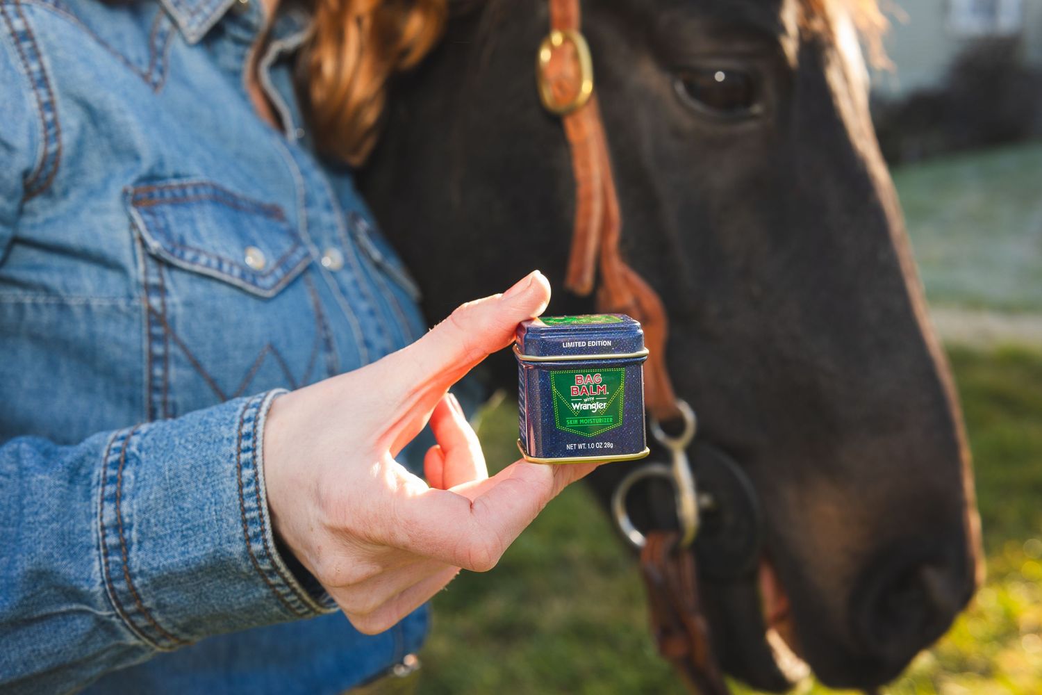Woman in denim jacket holding Bag Balm x Wrangler tin with brown horse in background.