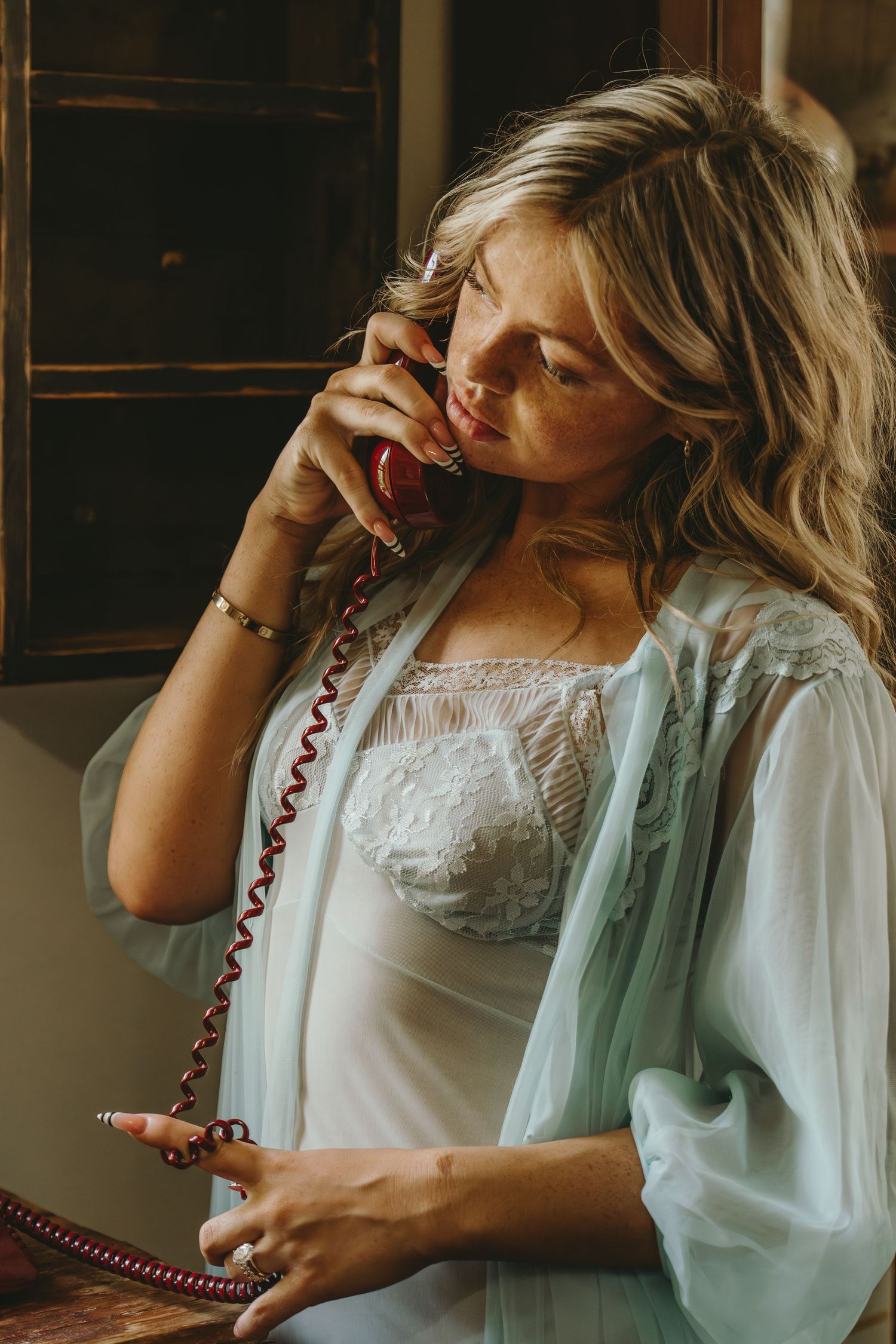 Blonde woman in sheer blue robe talking on a vintage red rotary telephone indoors.