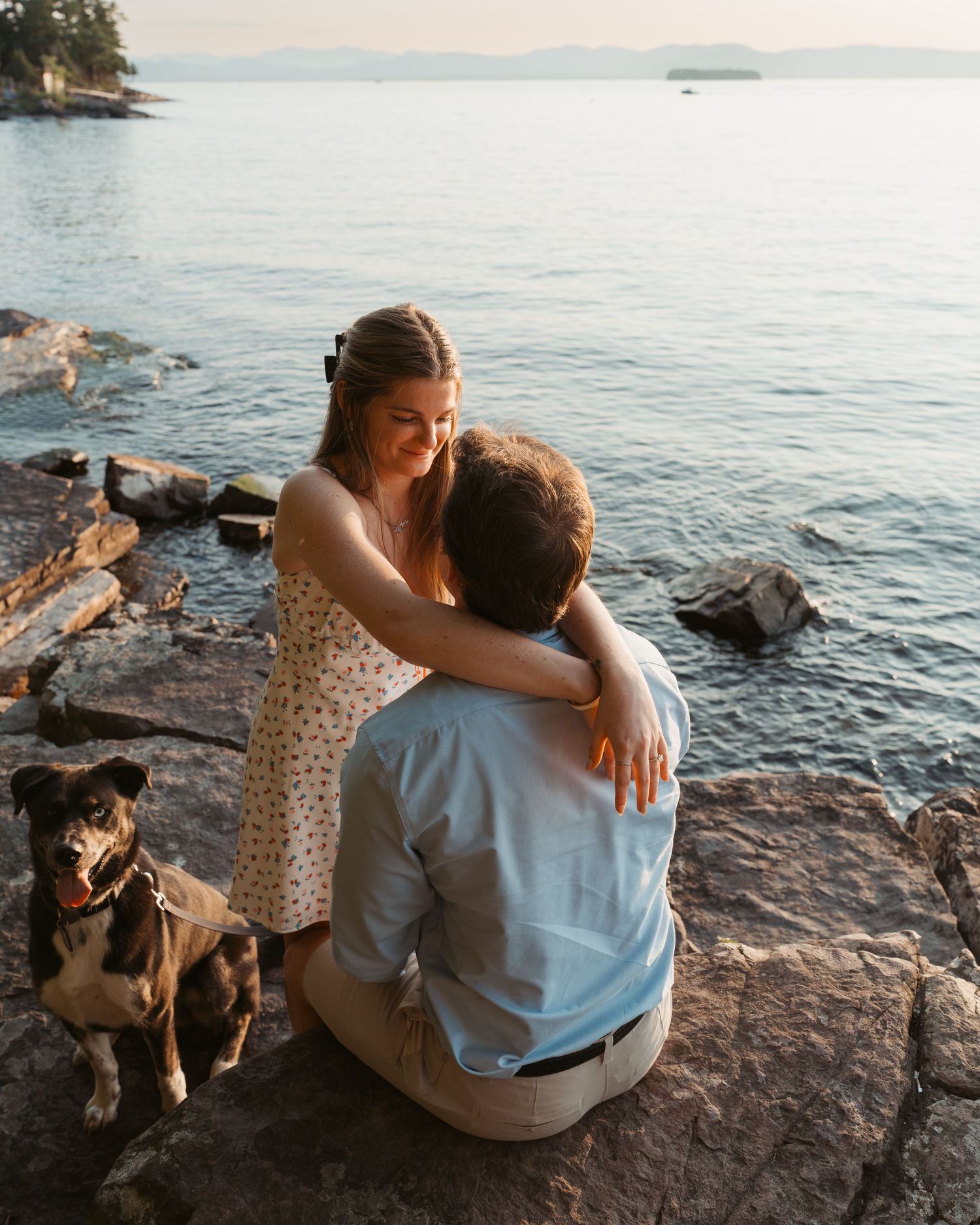 Couple embracing on rocky lakeside at sunset with a black and white dog nearby.