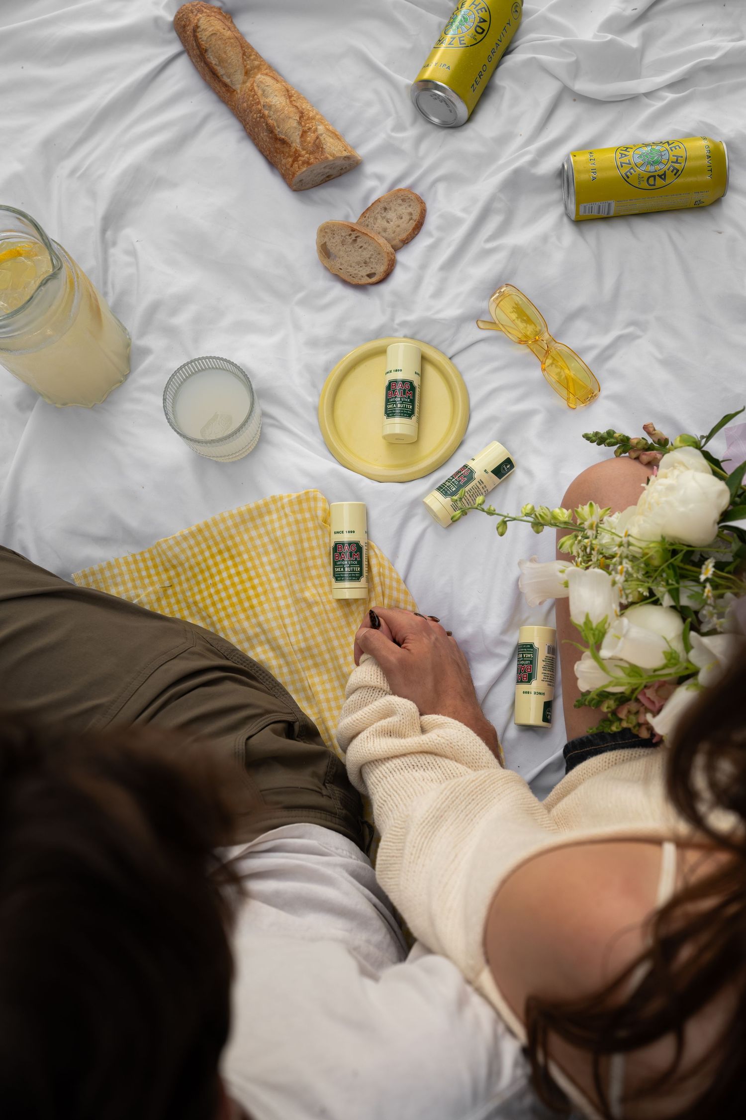Couple enjoying a picnic on white blanket with Bag Balm products, lemonade cans, bread, and white flowers.
