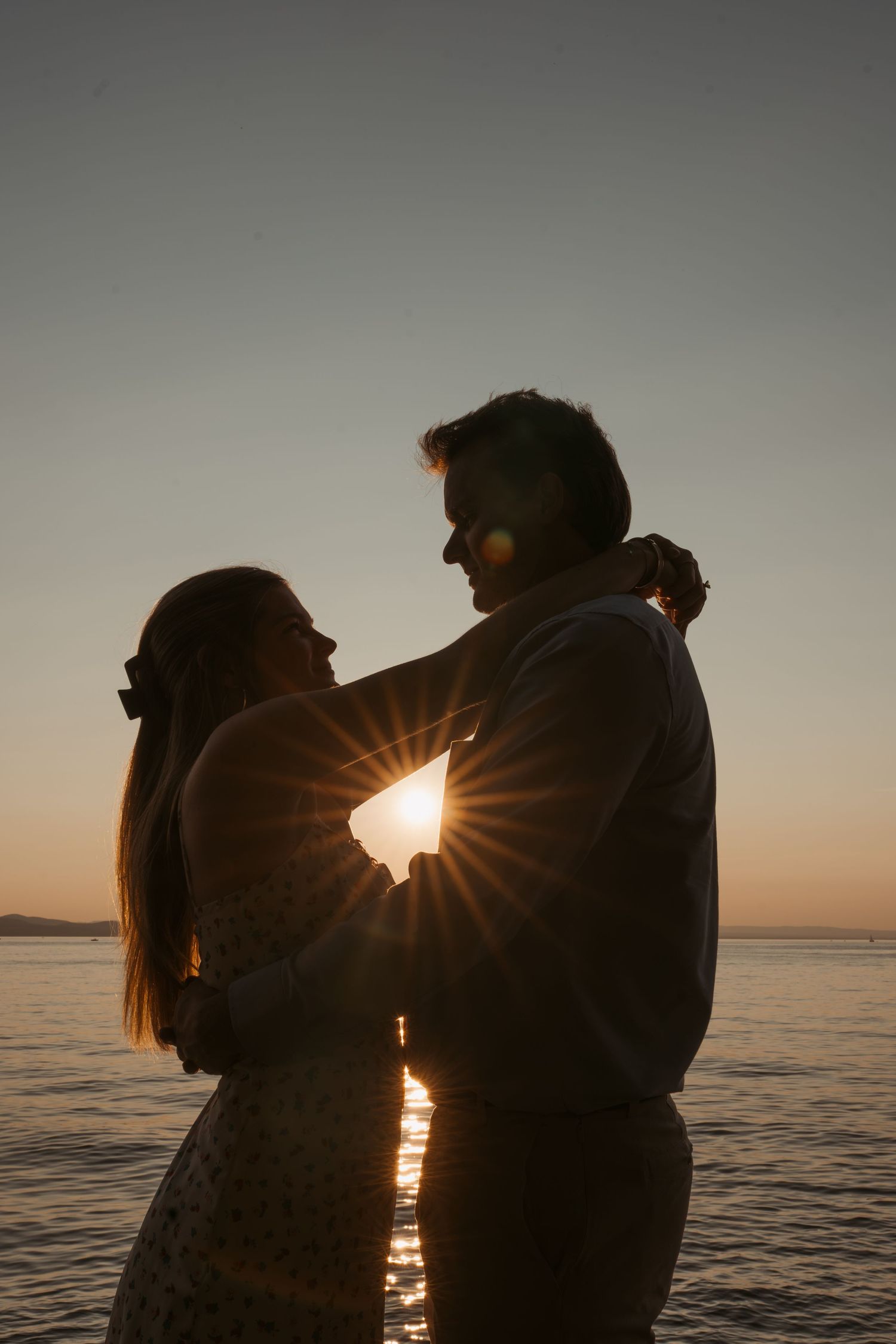 Romantic silhouette of couple embracing at sunset by the ocean with sunburst effect.