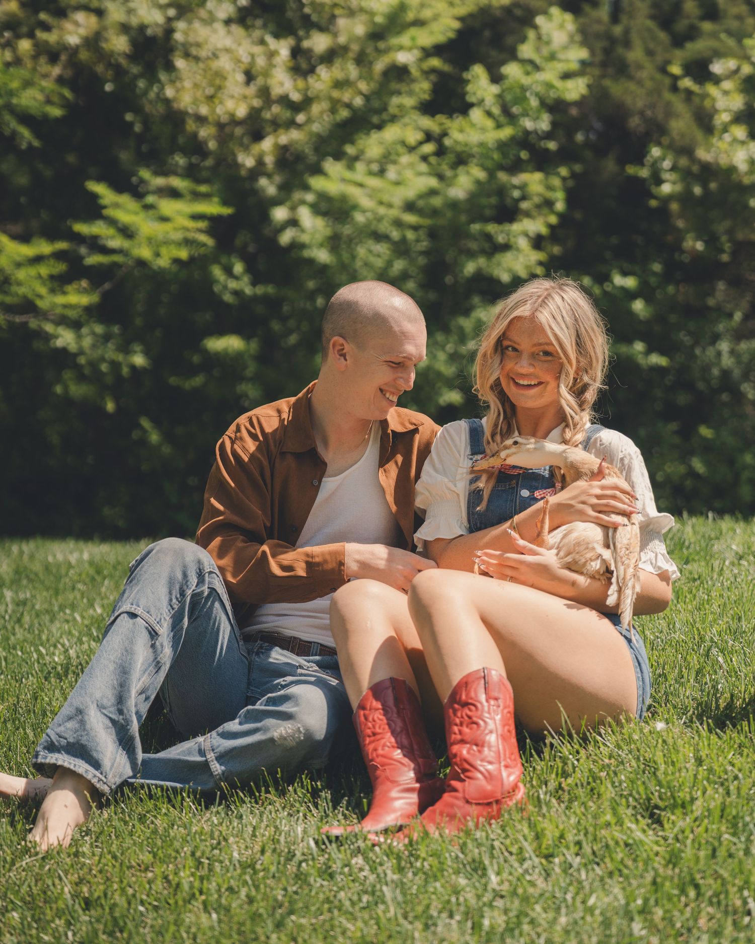 A happy couple sits on green grass in sunlight, holding a small duck, dressed in casual country-style outfits.