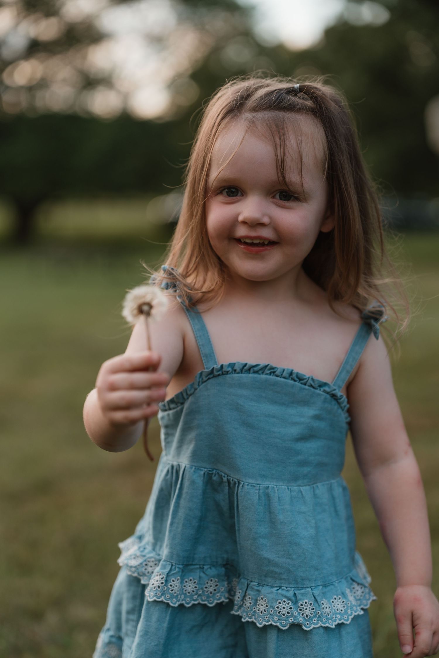 Smiling toddler girl in a blue lace dress holding a dandelion in a sunny green park.
