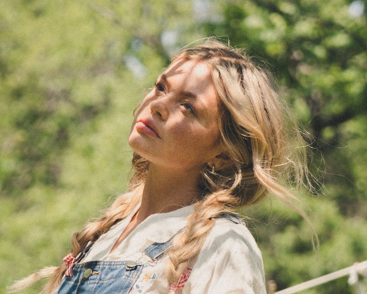 Close-up of a blonde woman with braids looking up in natural sunlight outdoors.