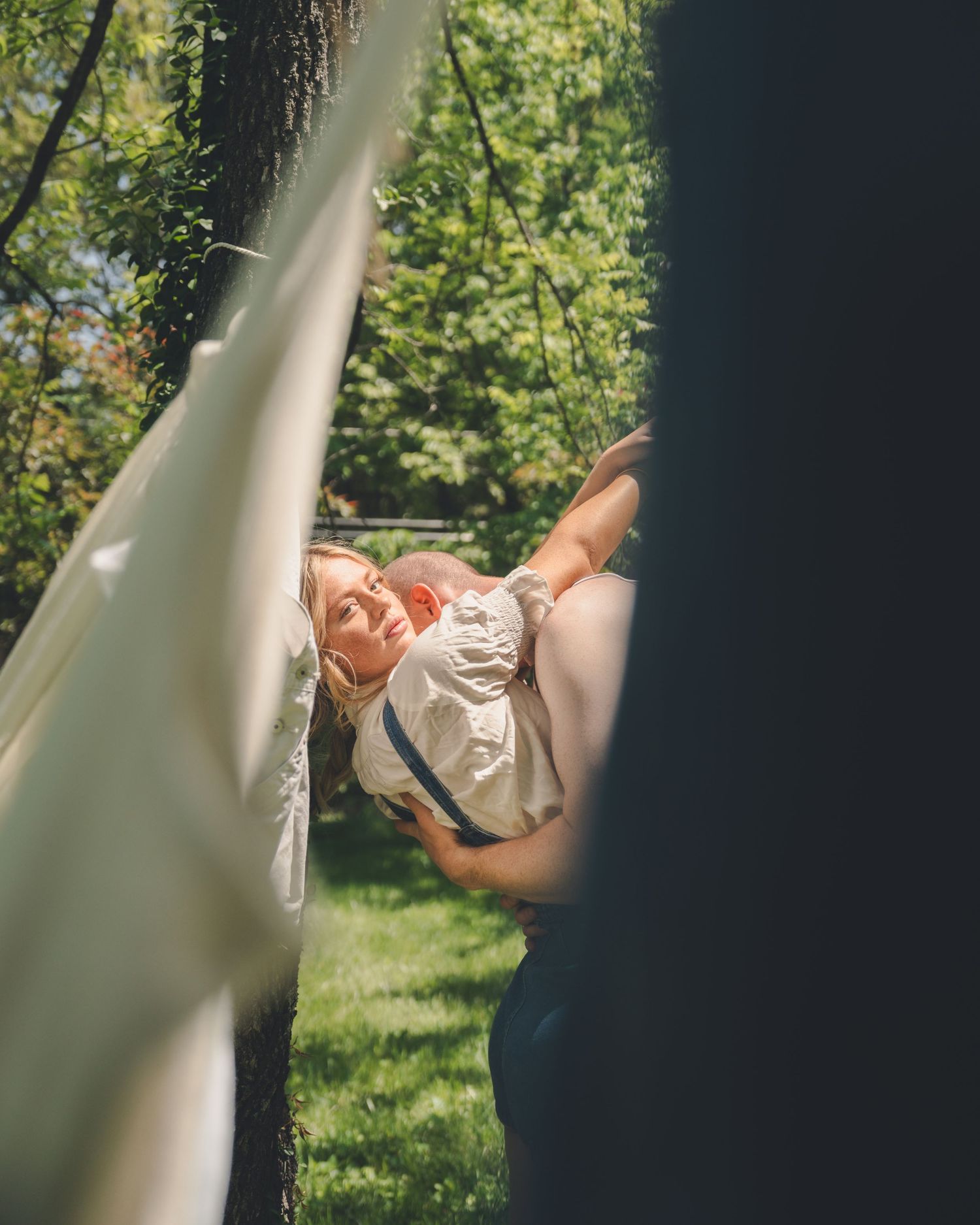 Woman being lifted by partner in a sunny green garden, captured through hanging fabric.