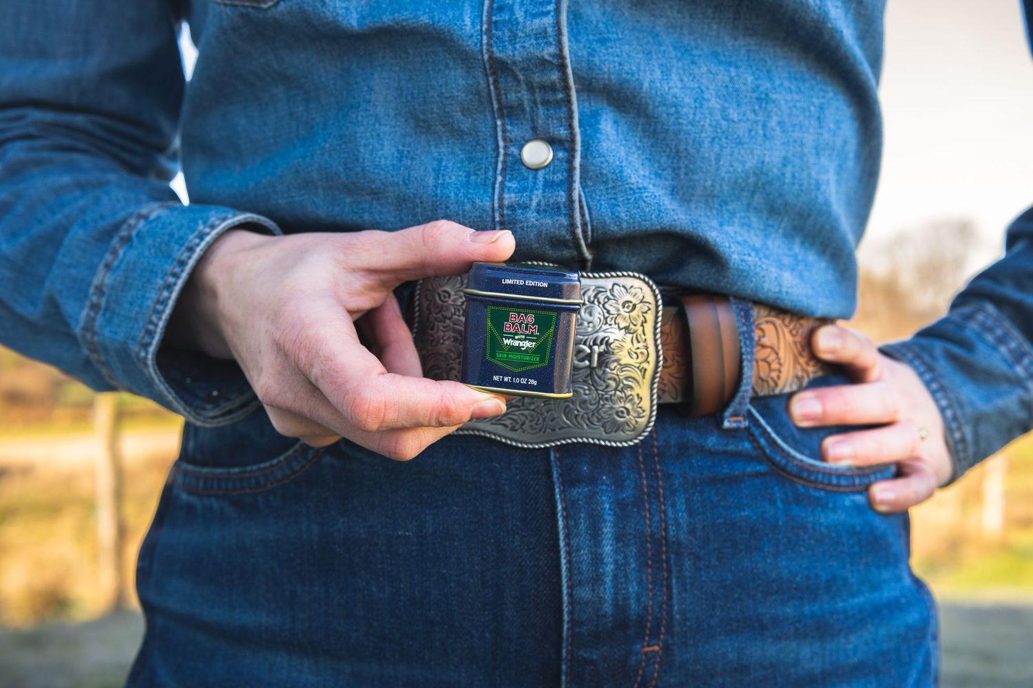 Person in denim shirt and Wrangler belt holding Bag Balm x Wrangler limited edition tin outdoors.