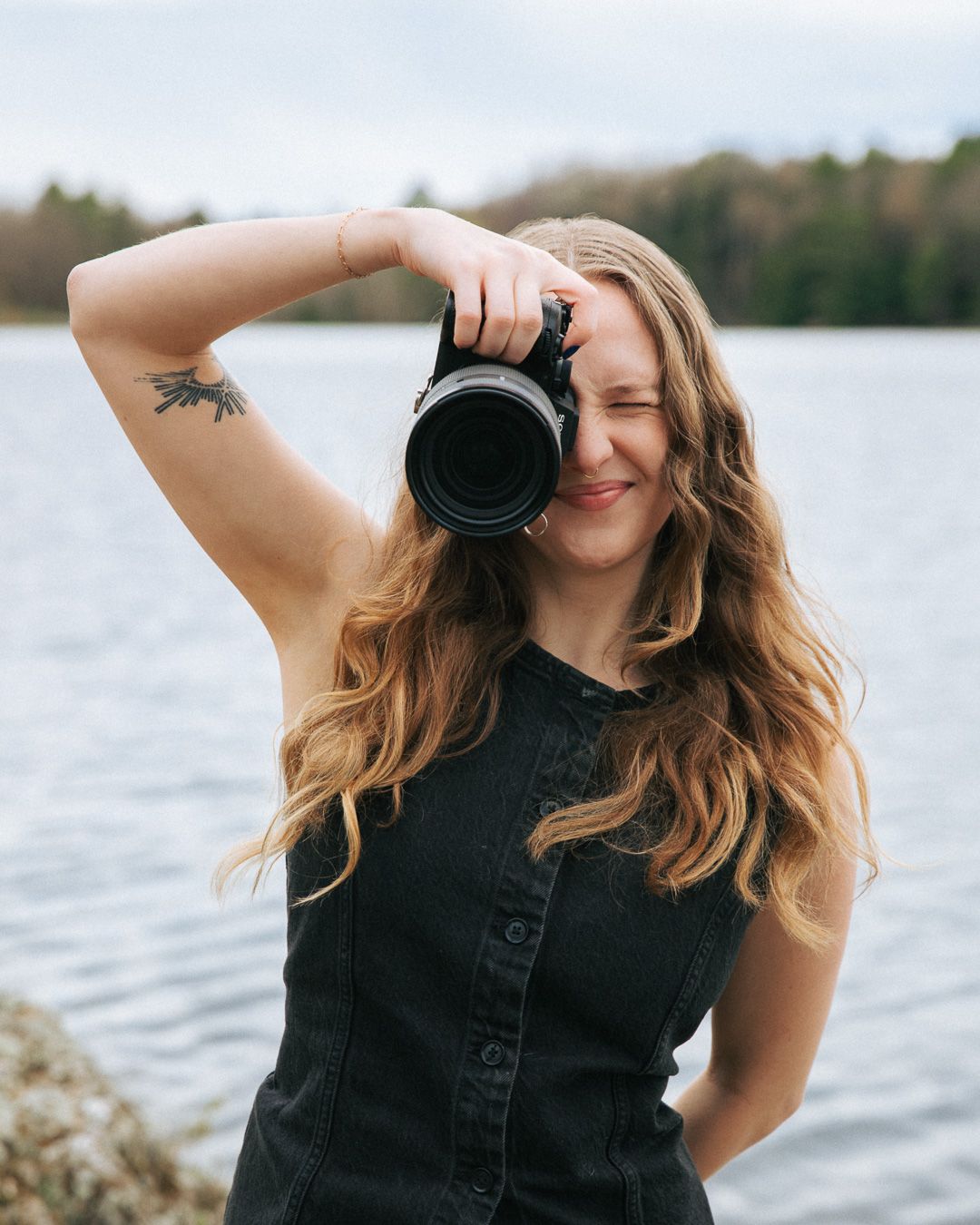 Young woman with long wavy hair holds camera to her eye, smiling outdoors near a lake.