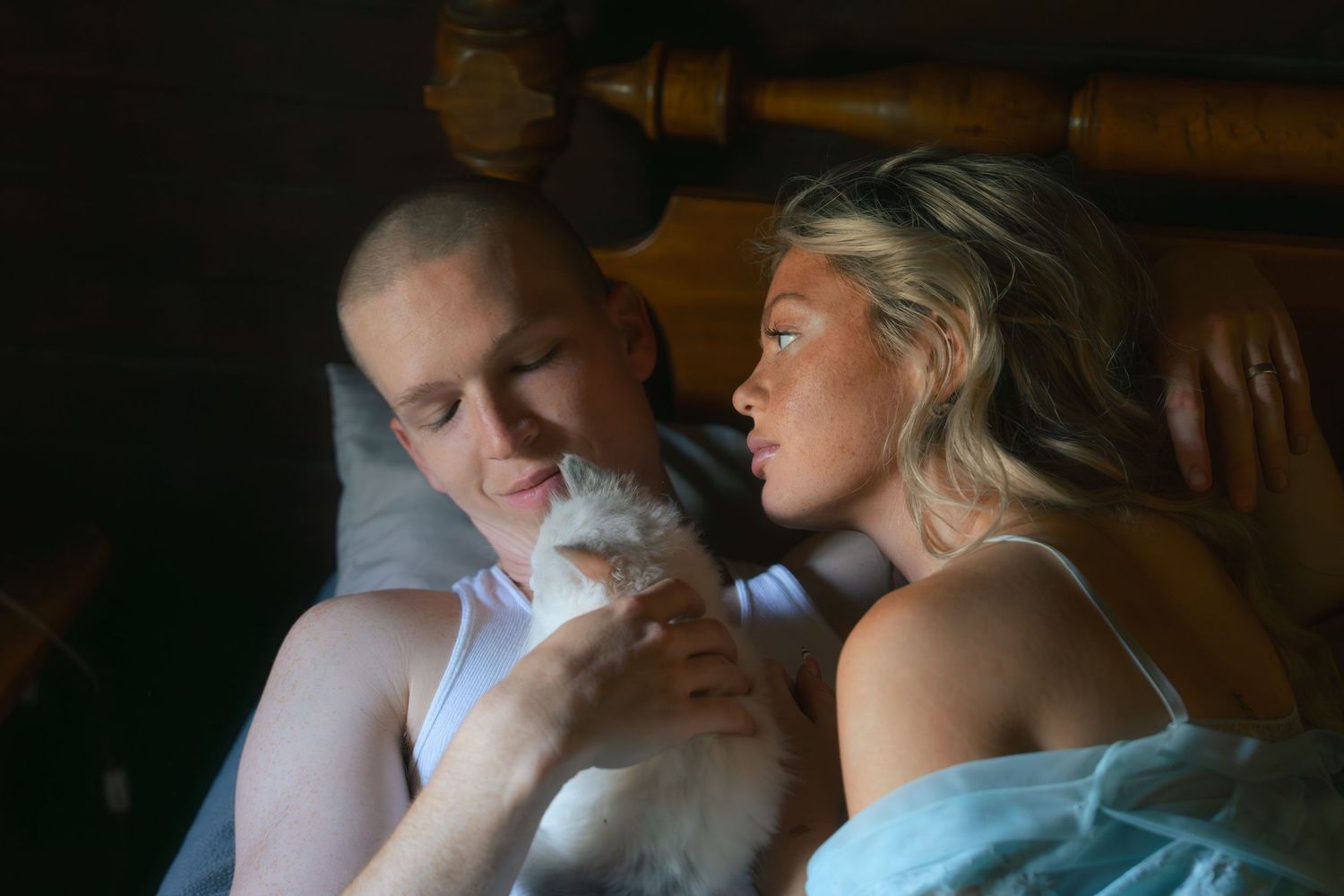 A couple relaxes together in a cozy dark room, cuddling with a small white cat between them.