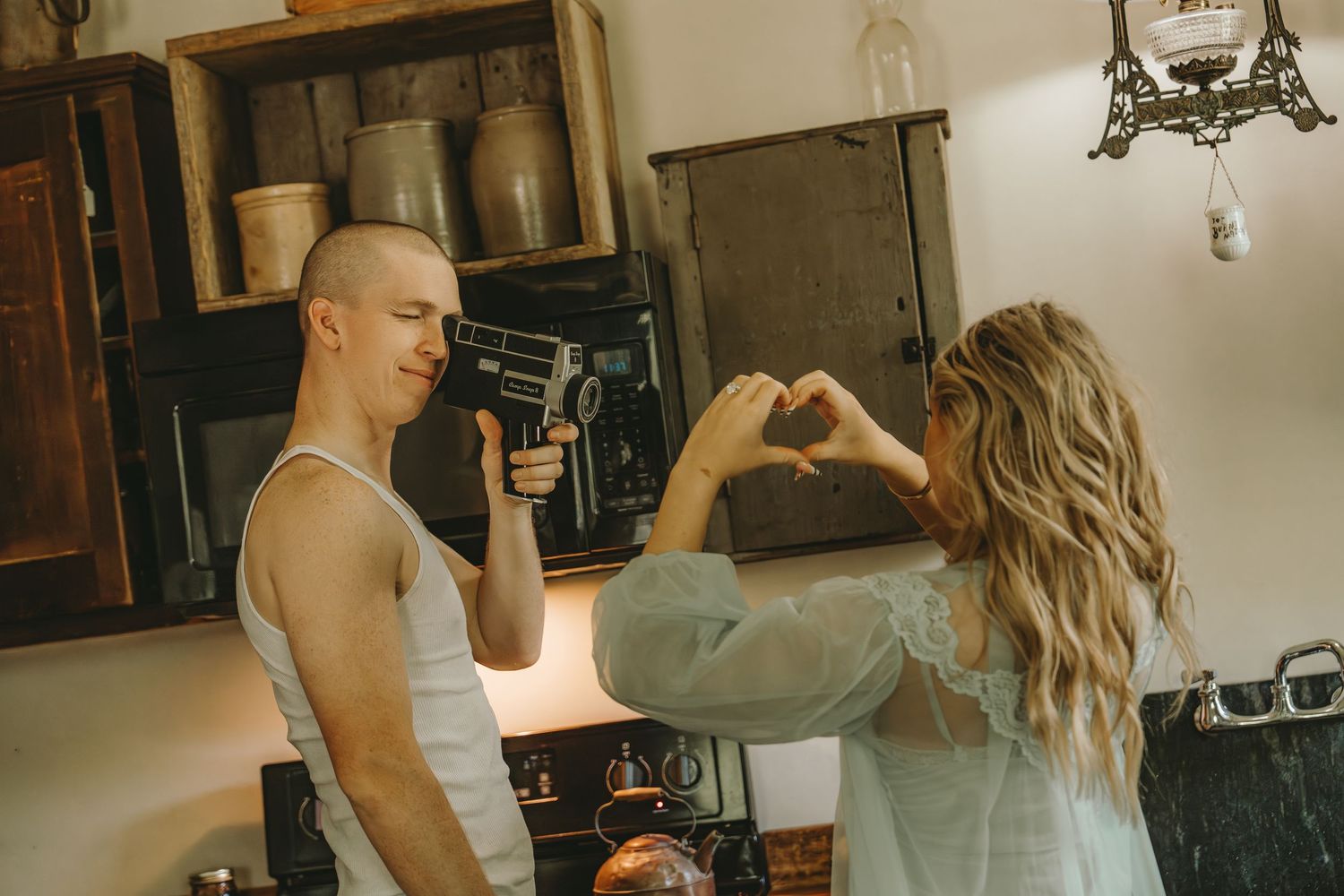 Man filming woman with vintage camera as she makes a heart shape with her hands in a rustic kitchen.