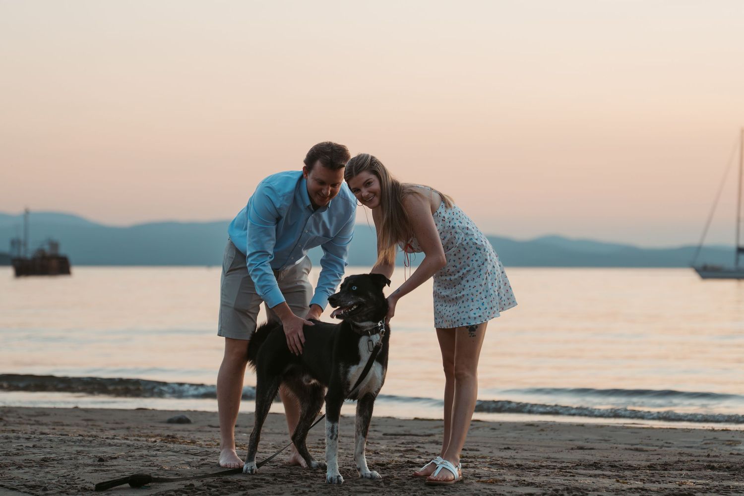 Couple petting their black dog together on a sandy beach at sunset.