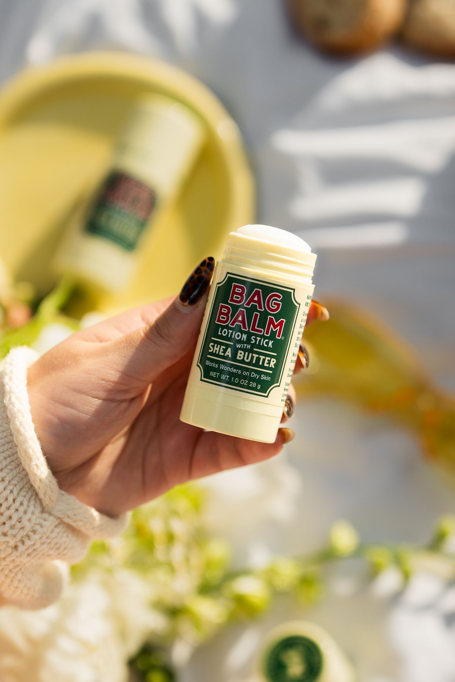 Hand holding Bag Balm Shea Butter lotion stick surrounded by spring flowers.