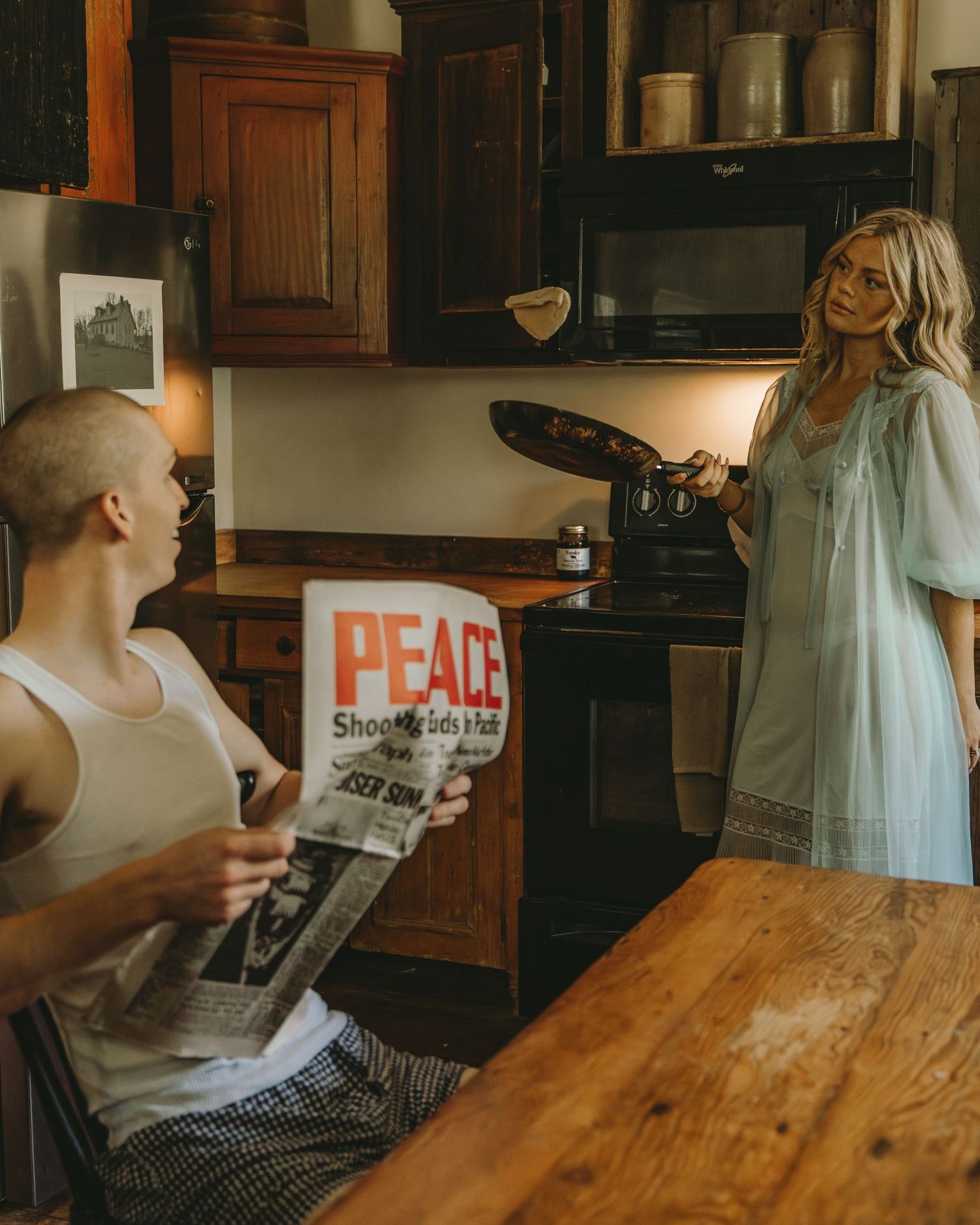 A blonde woman in a blue robe flips a pancake in a rustic kitchen while a man reads a peace newspaper.
