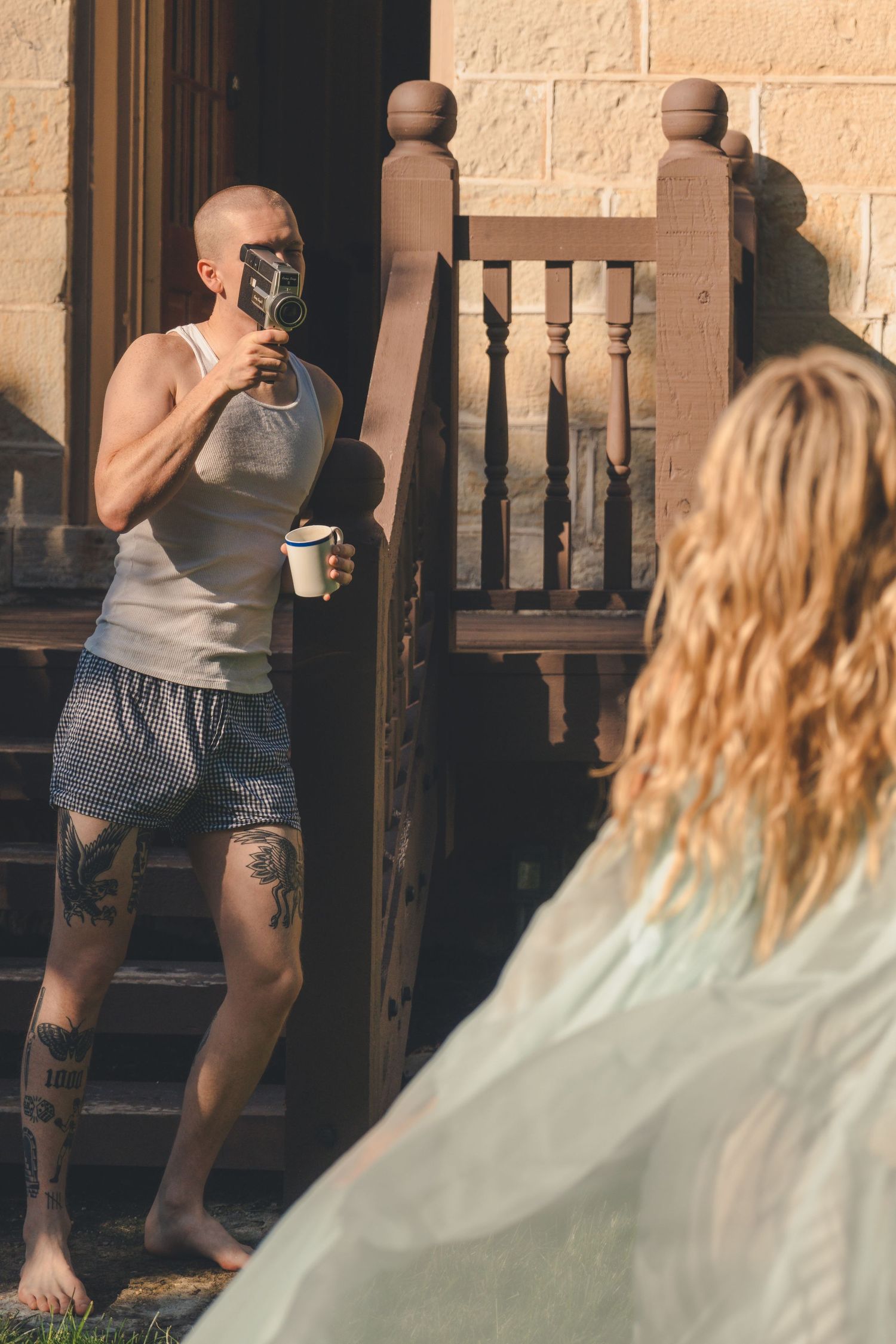 A tattooed man in a tank top holds a coffee mug and a plant on a sunny wooden porch steps.