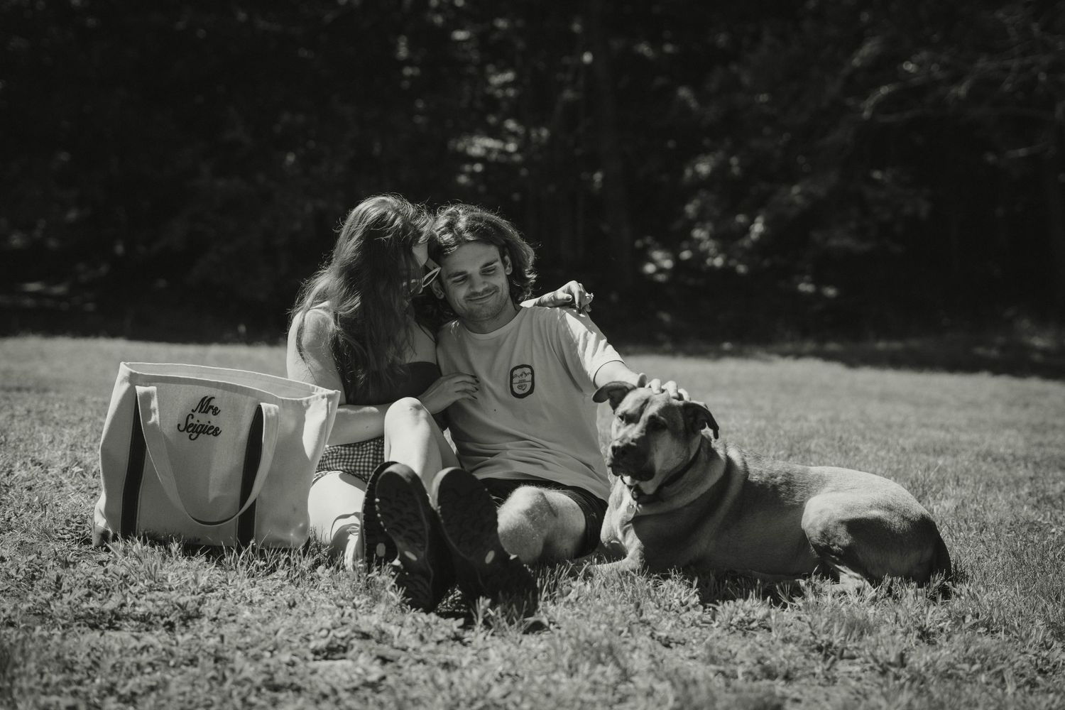 Black and white photo of couple relaxing on grass with large dog lying beside them.