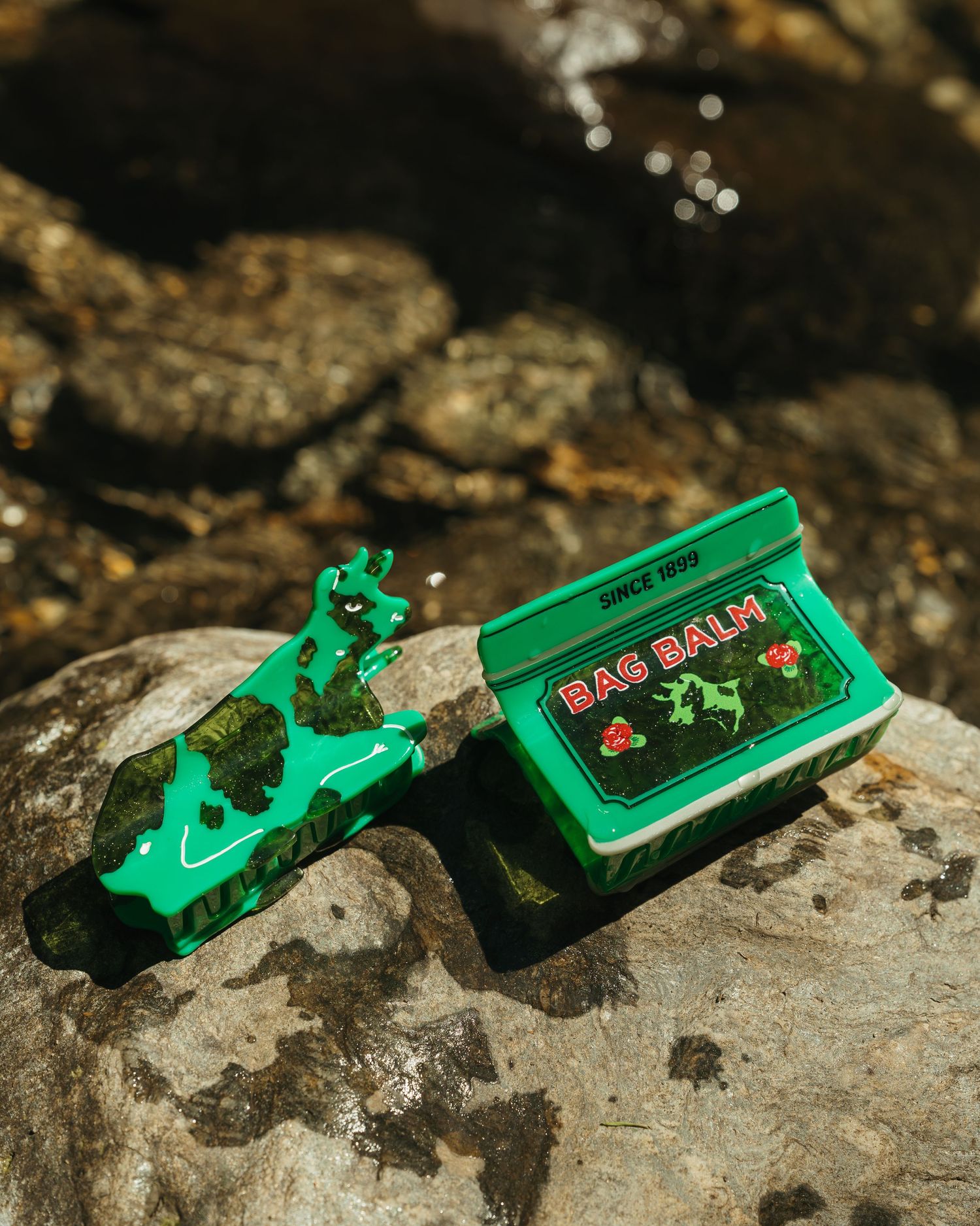 Green Bag Balm tin and hair clip accessory displayed on rocks in sunlight.