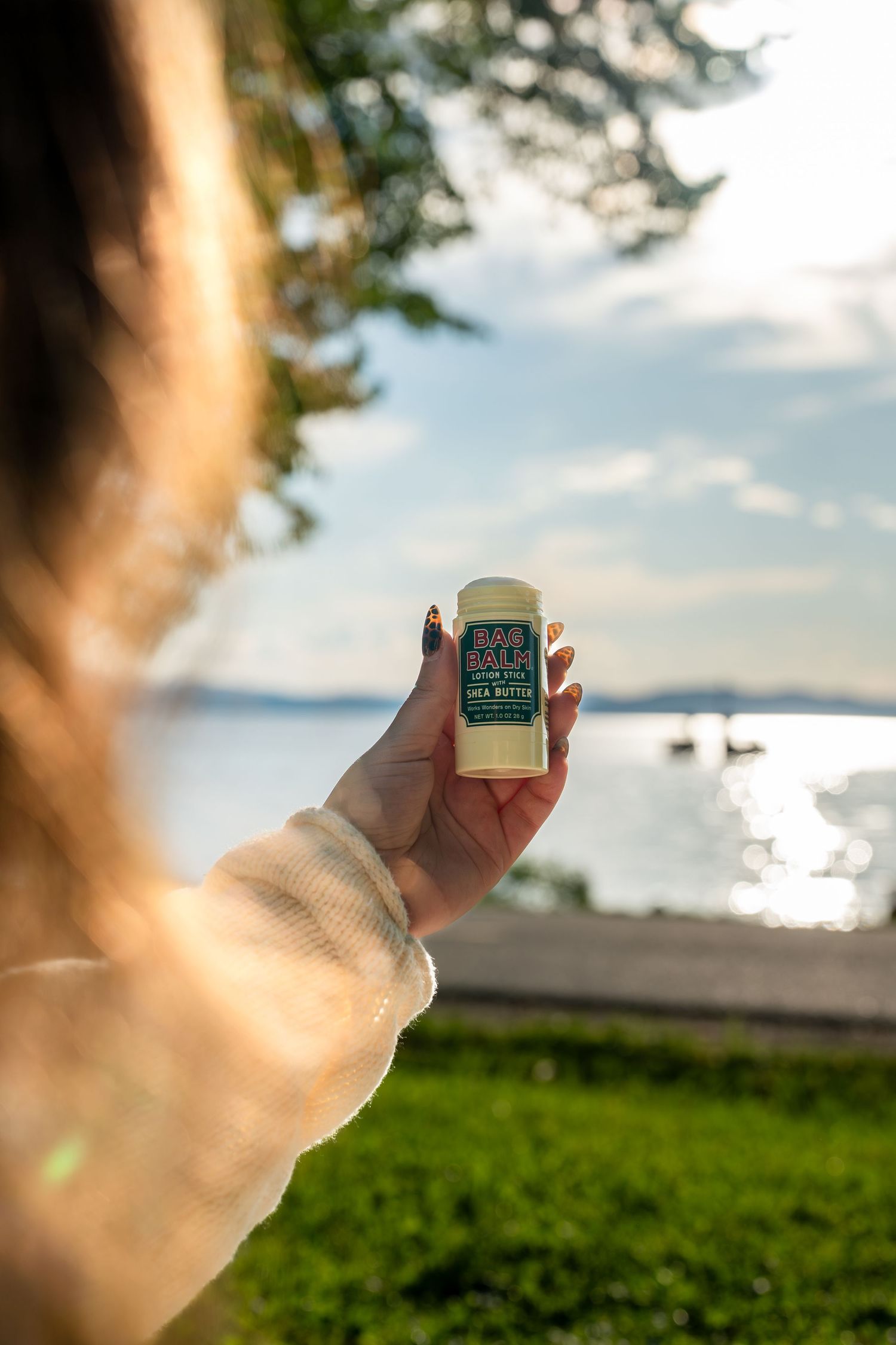 Woman holding Bag Balm stick with scenic lake and sunset view in background.