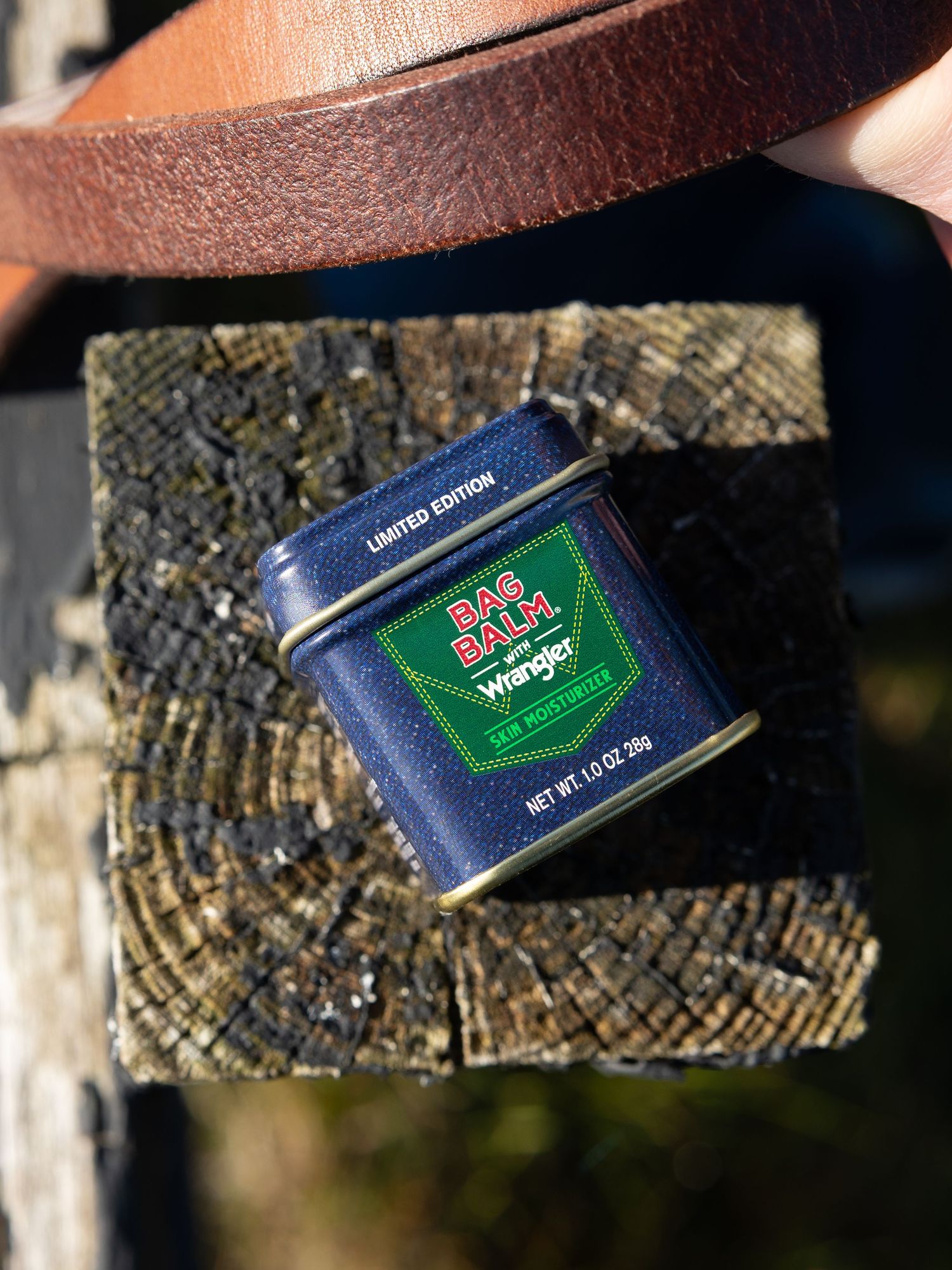 Limited edition Bag Balm x Wrangler skin moisturizer tin resting on rustic wooden post outdoors.