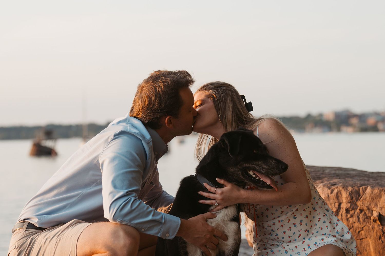 Couple kissing while holding their black dog on a rocky waterfront at dusk.