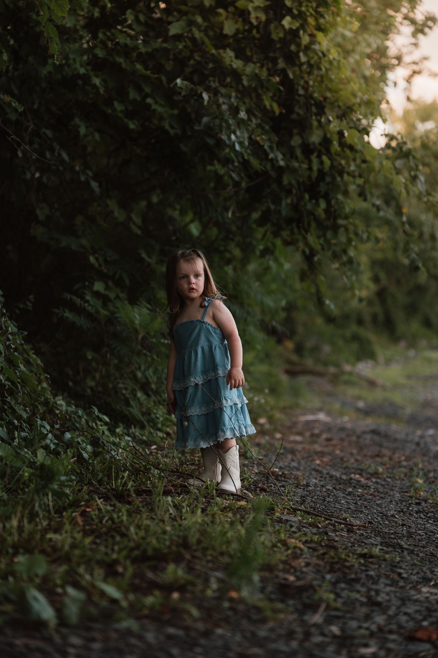 Young girl in a teal dress standing alone on a gravel path surrounded by lush green foliage.