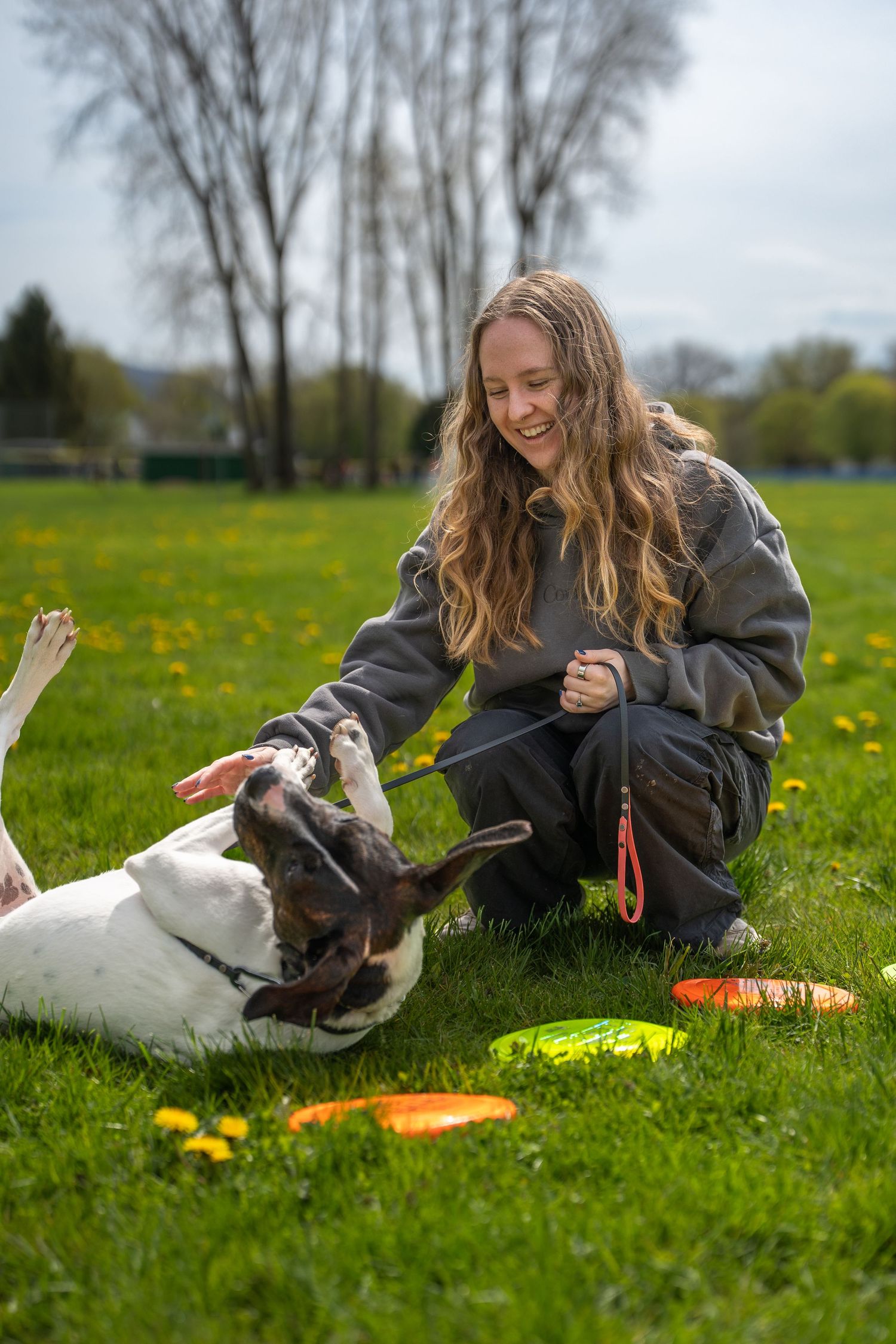 Young woman laughing while playing with her dog on green grass in a park, with colorful frisbees nearby.