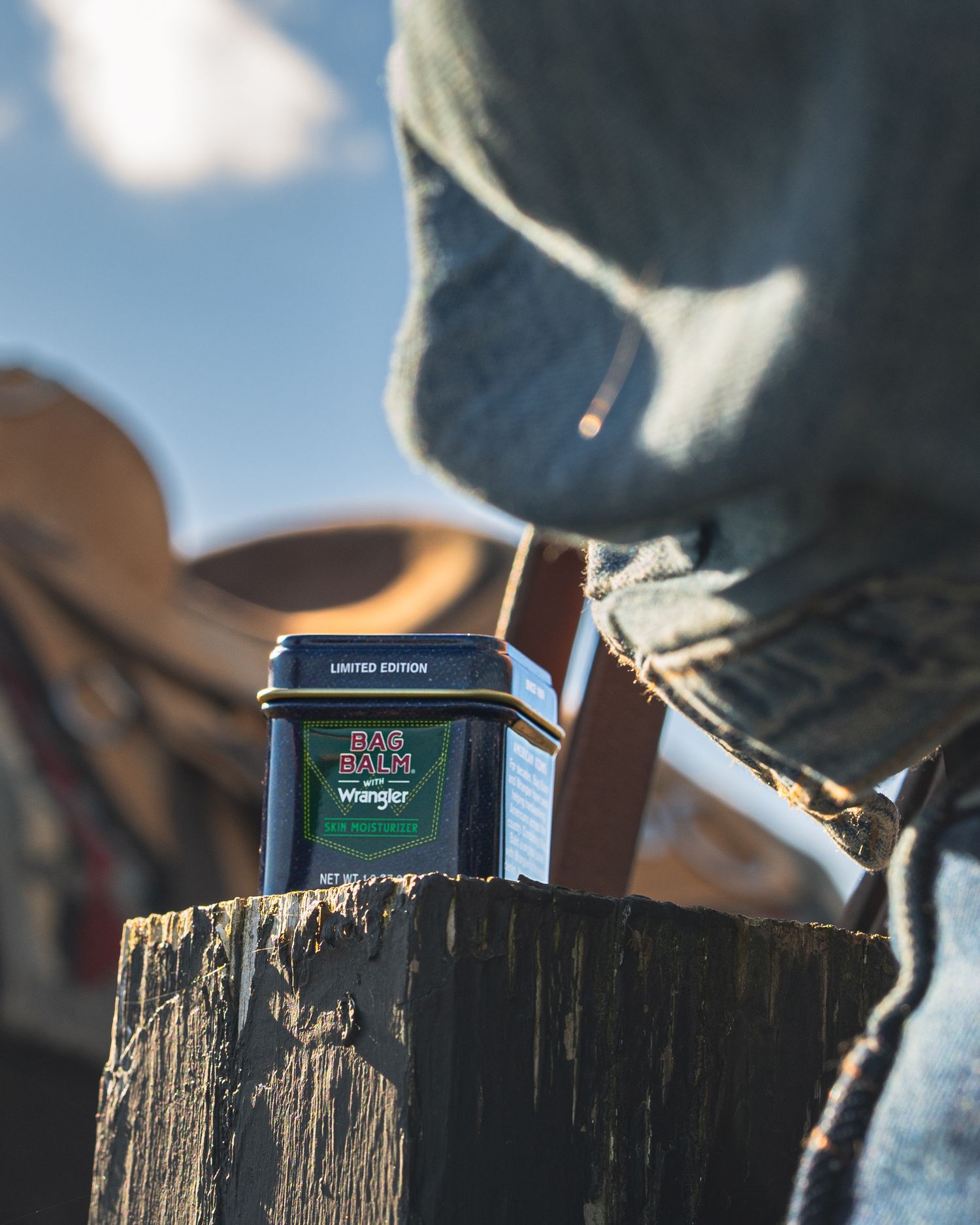 Limited edition Bag Balm Wrangler tin on wooden post with saddle and hooded person in background.