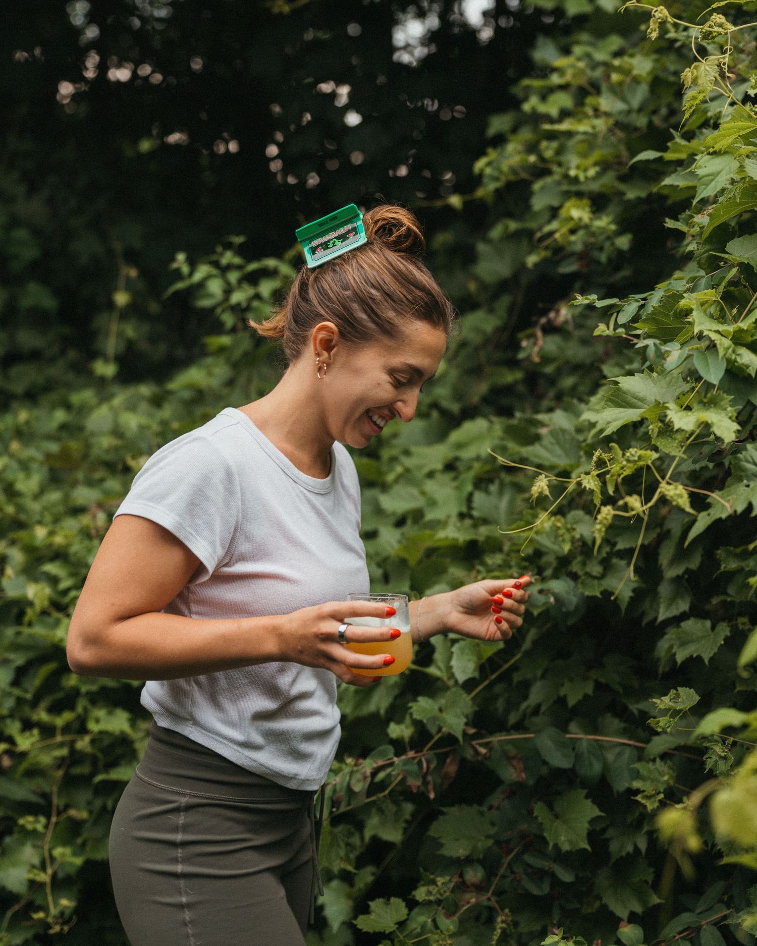 Smiling woman holding a drink while picking hops from green vines outdoors.