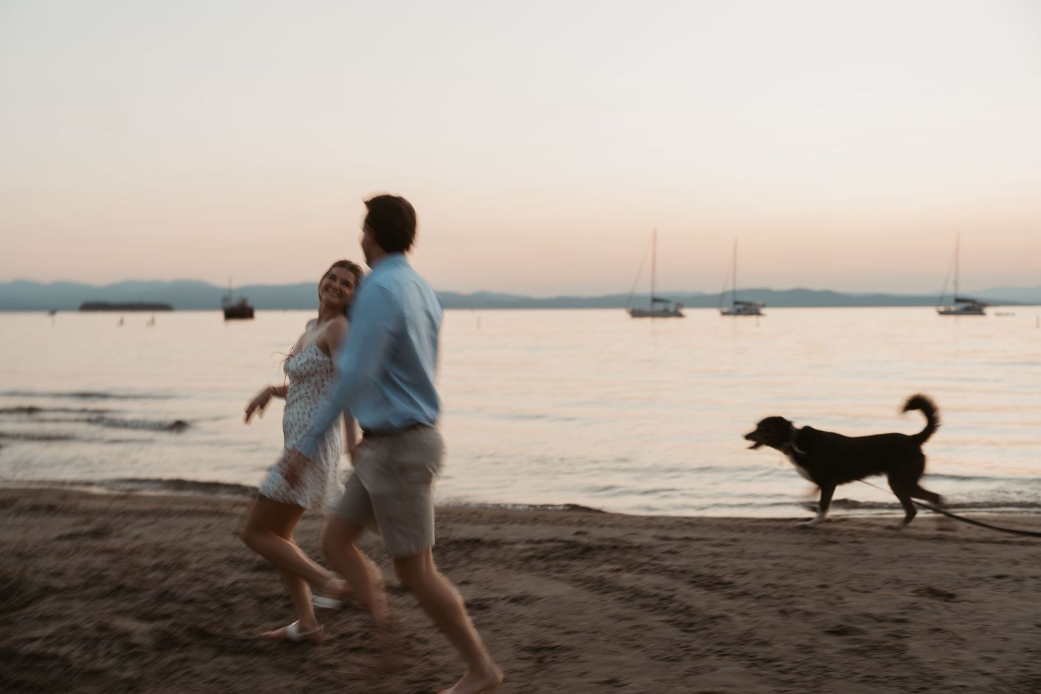 Couple running on beach at sunset with dog playing near calm water and sailboats in background.