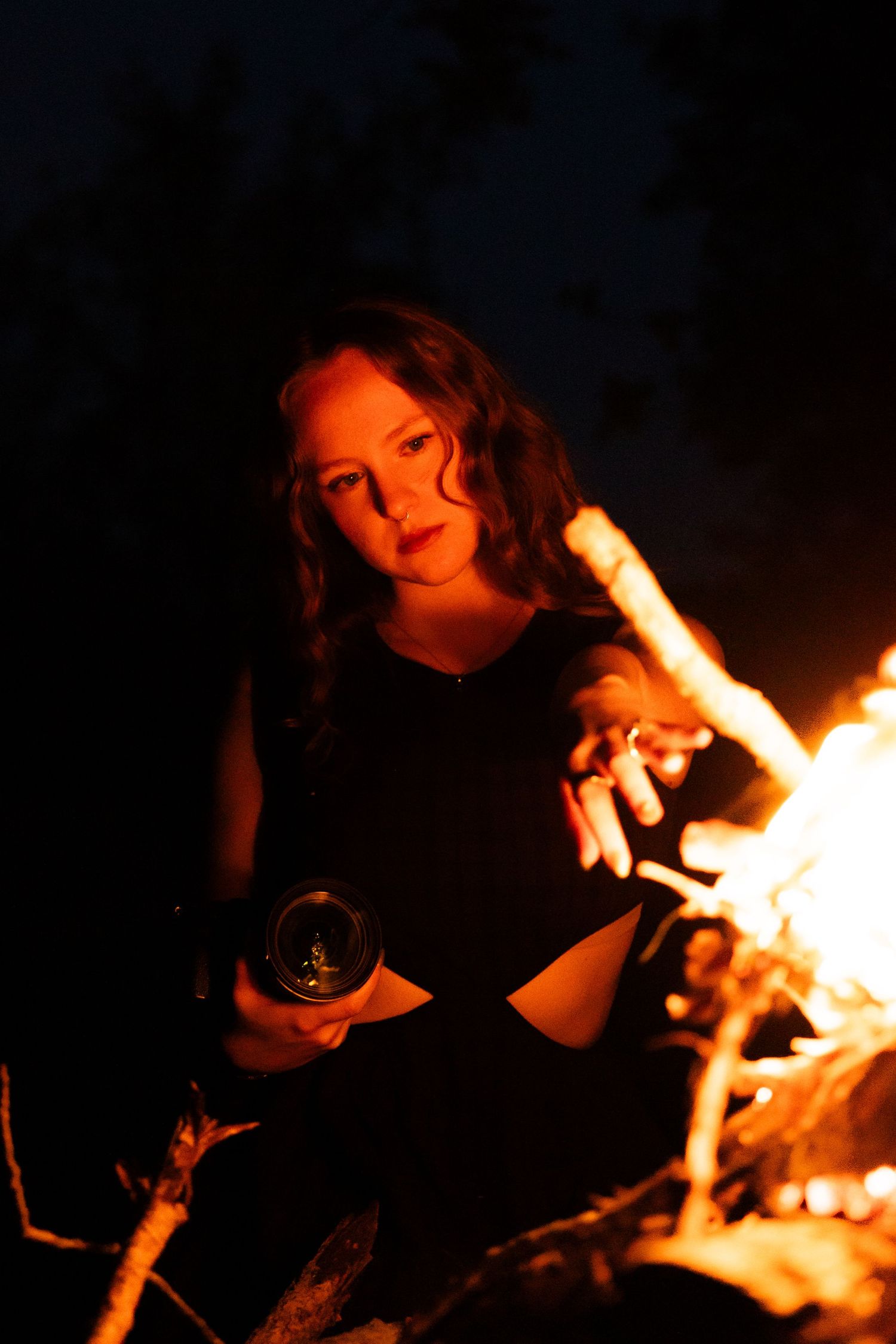 Woman in black outfit sitting by campfire at night holding a camera.