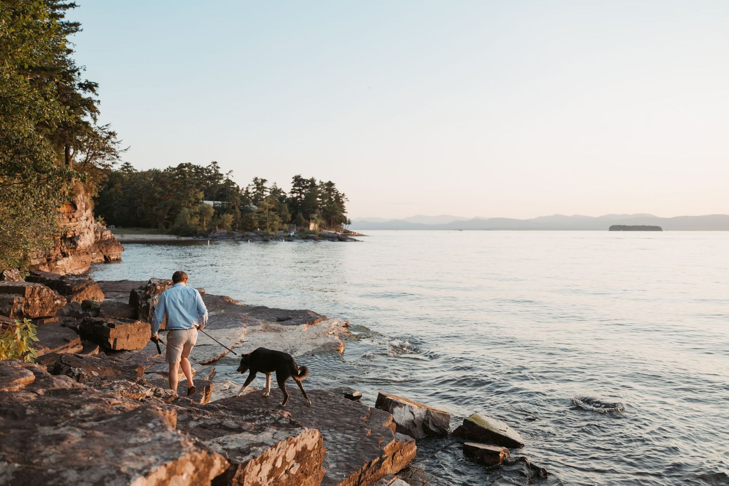 Person walking a black dog along rocky lakeside shore surrounded by trees at golden hour.