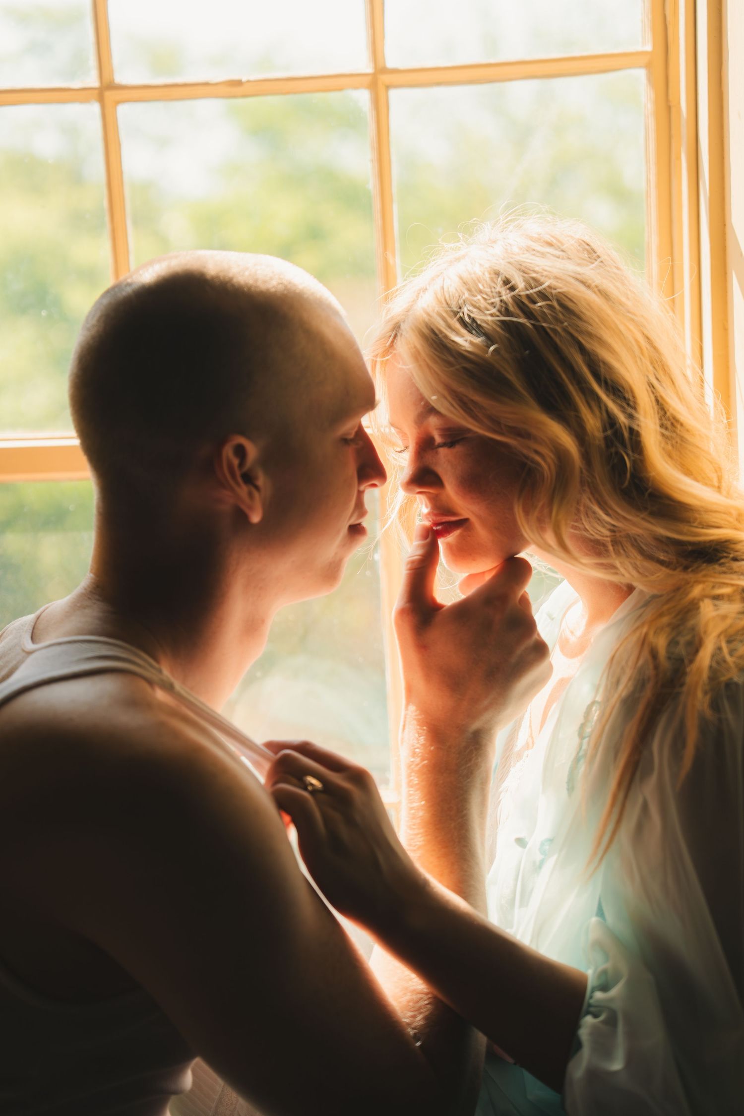 A couple shares a tender almost-kiss moment bathed in warm golden sunlight by a large window.