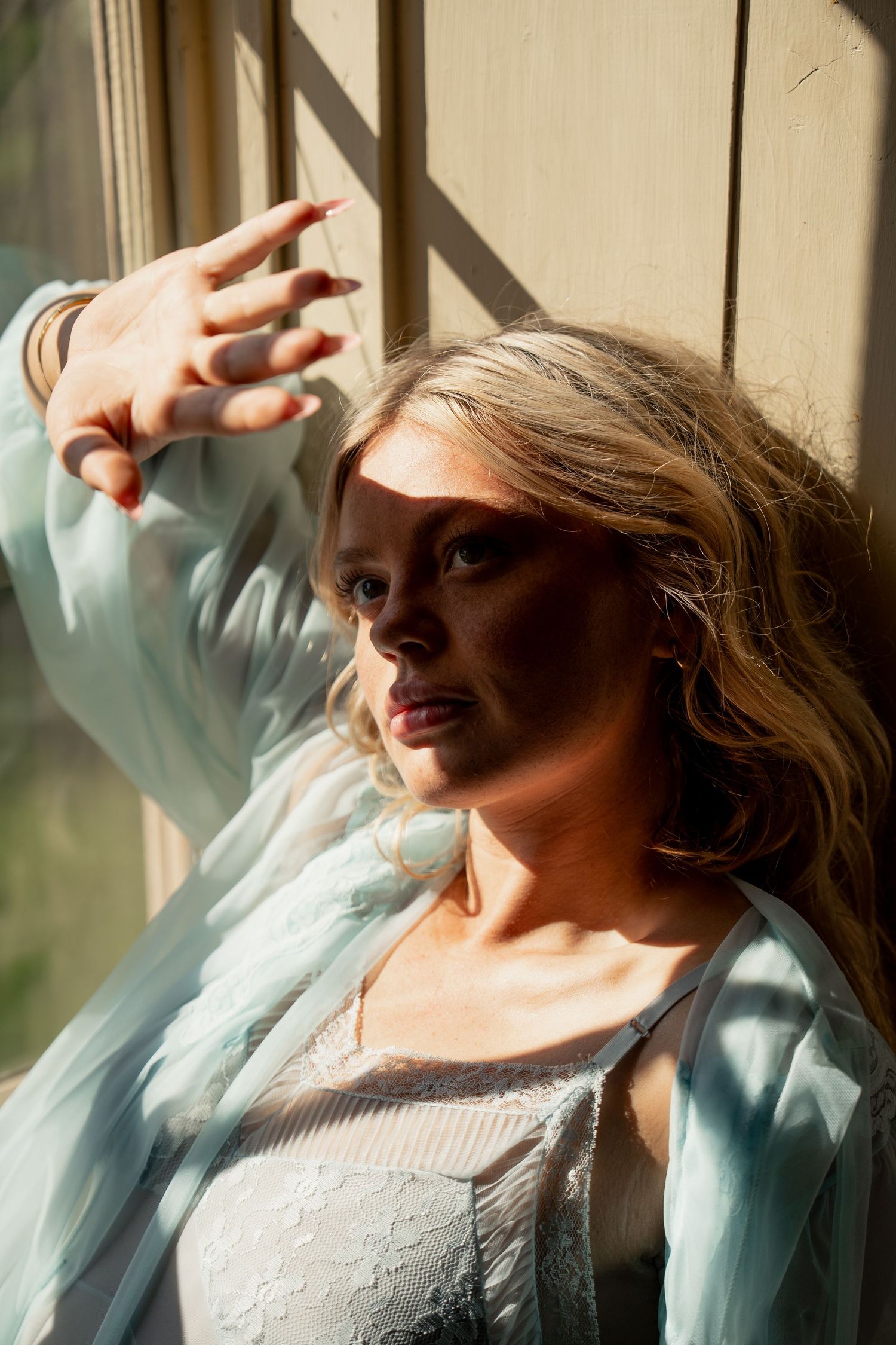 Blonde woman in sheer blue robe shielding her face from sunlight near a window.