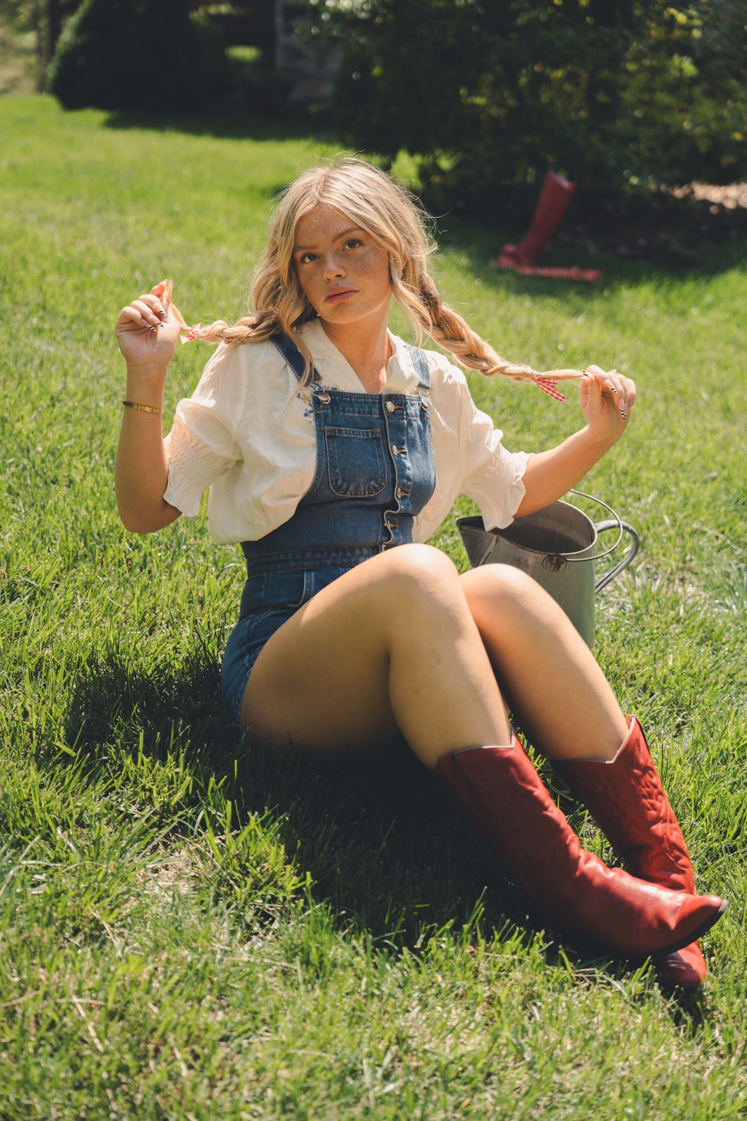 Blonde woman in denim overalls and red boots sitting on grass holding braids.